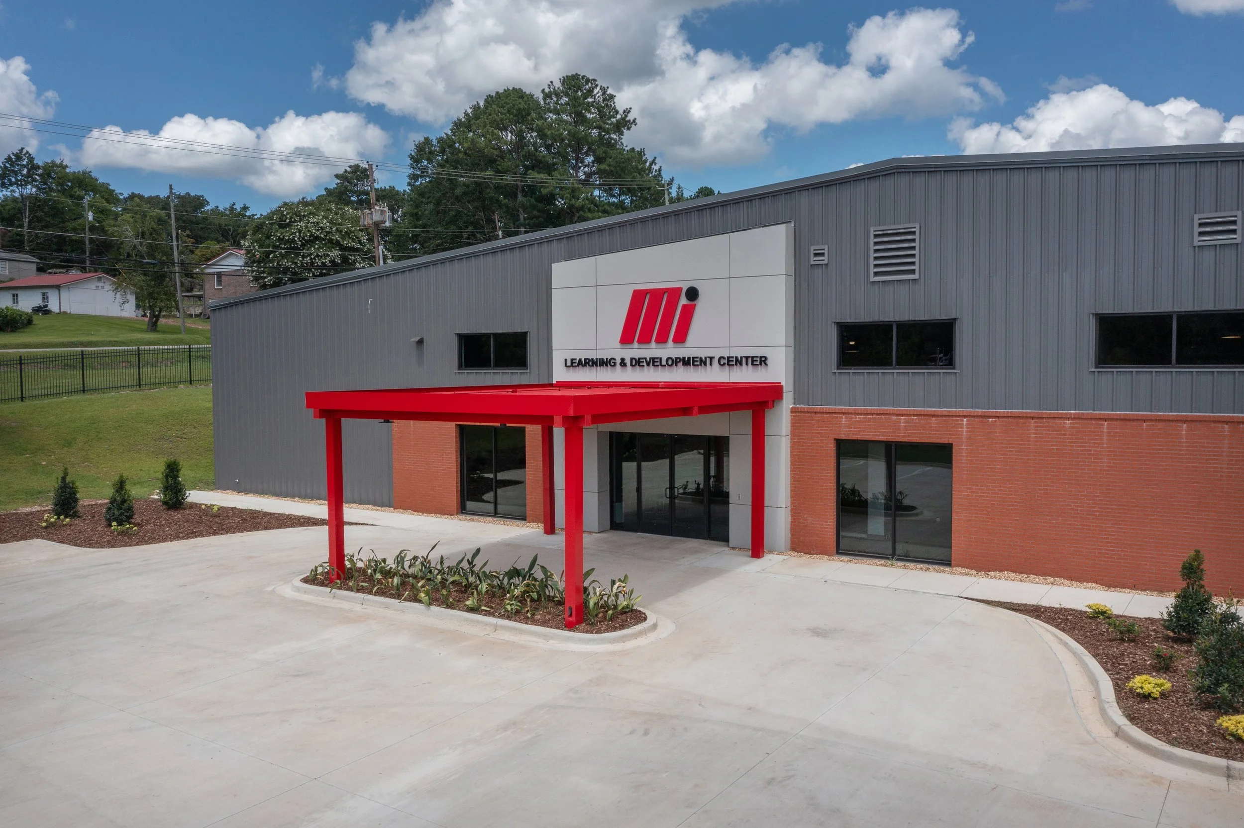 Exterior view of a Training Facility for Motion Industries located in Birmingham AL with gray metal siding, brick walls, and a red entrance canopy, surrounded by a concrete driveway and landscaped plants.  project by B Group Architecture, Inc.