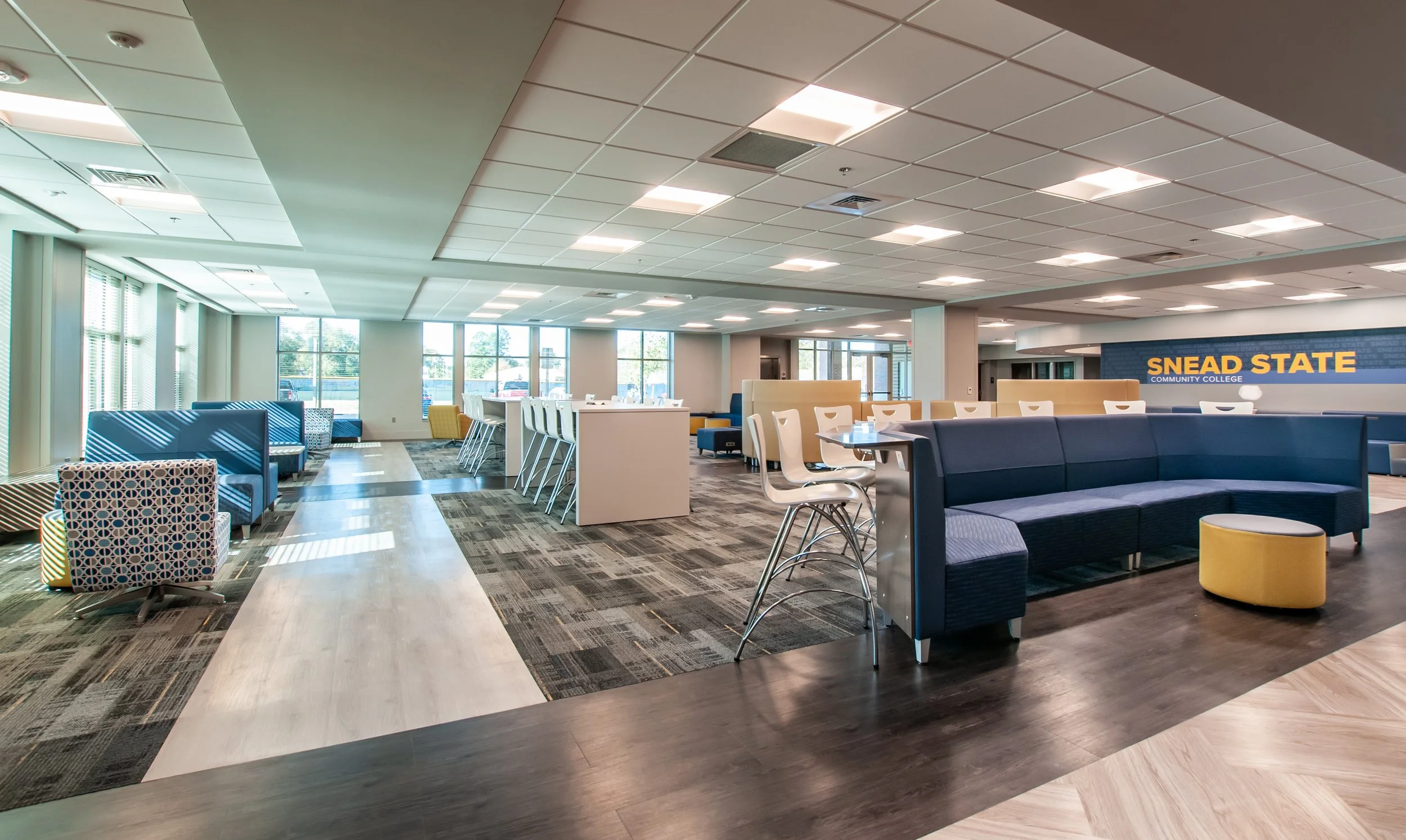 Modern community college lounge area with various seating options, including blue and patterned chairs, high tables with white chairs, and sofas. Large windows allow natural light, and a sign in the background reads 'Snead State Community College'.