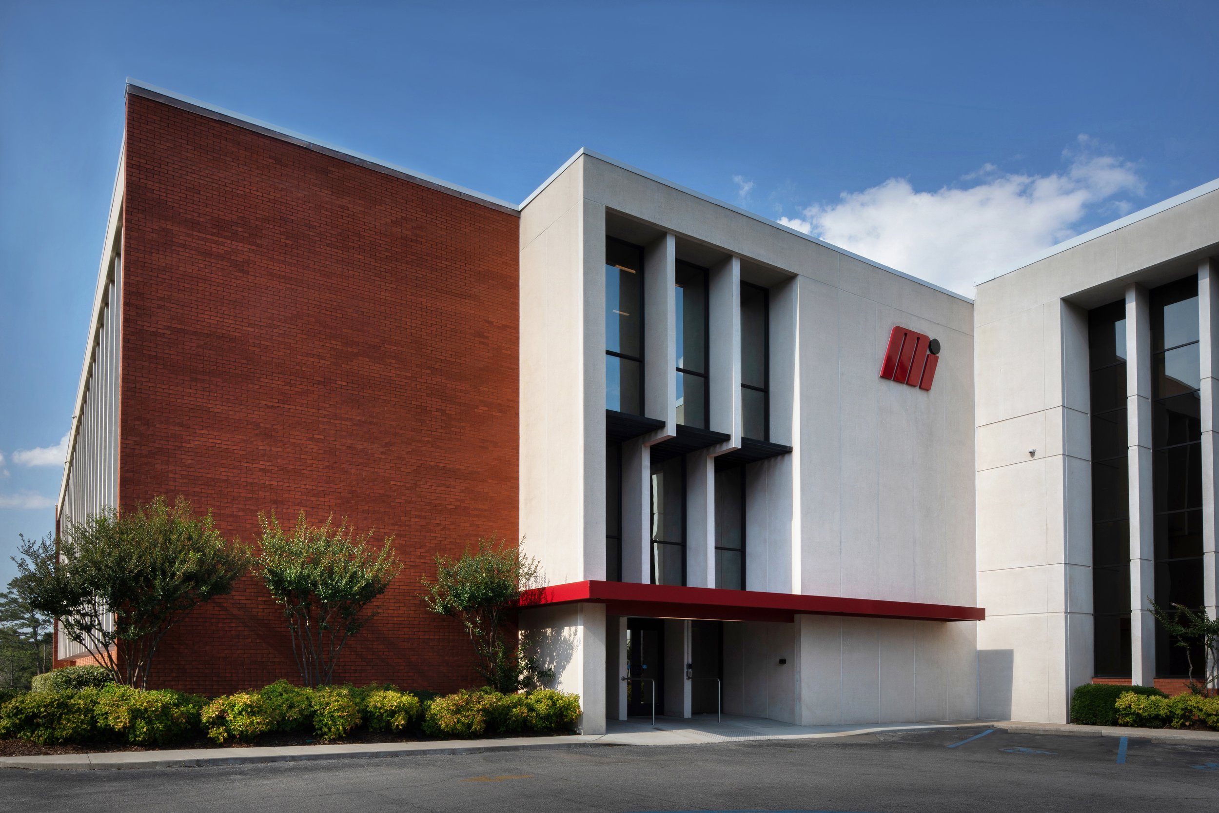 Modern office building with white and red brick facade, large windows, and a red logo on the upper right wall, surrounded by landscaping and a parking lot.