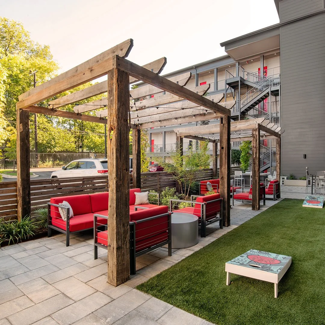 Outdoor patio area with red cushioned seating, and wooden pergola with a cornhole game for residents in a modern apartment complex. project by B Group Architecture, Inc.