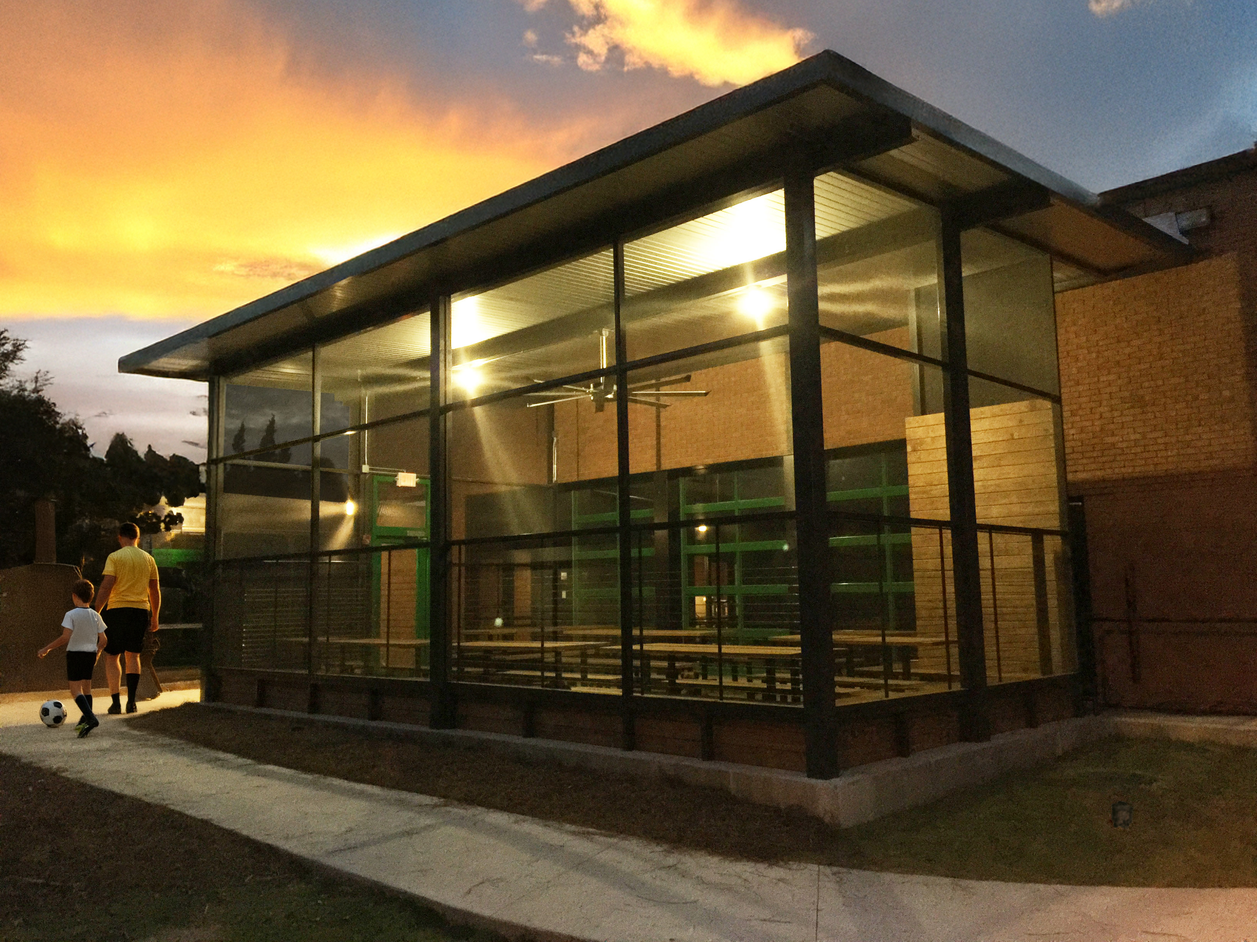 A modern glass building with a sloped roof at sunset. Inside, overhead lights are on, and outside, two people, one adult and one child, are walking along a sidewalk, with the child kicking a soccer ball.