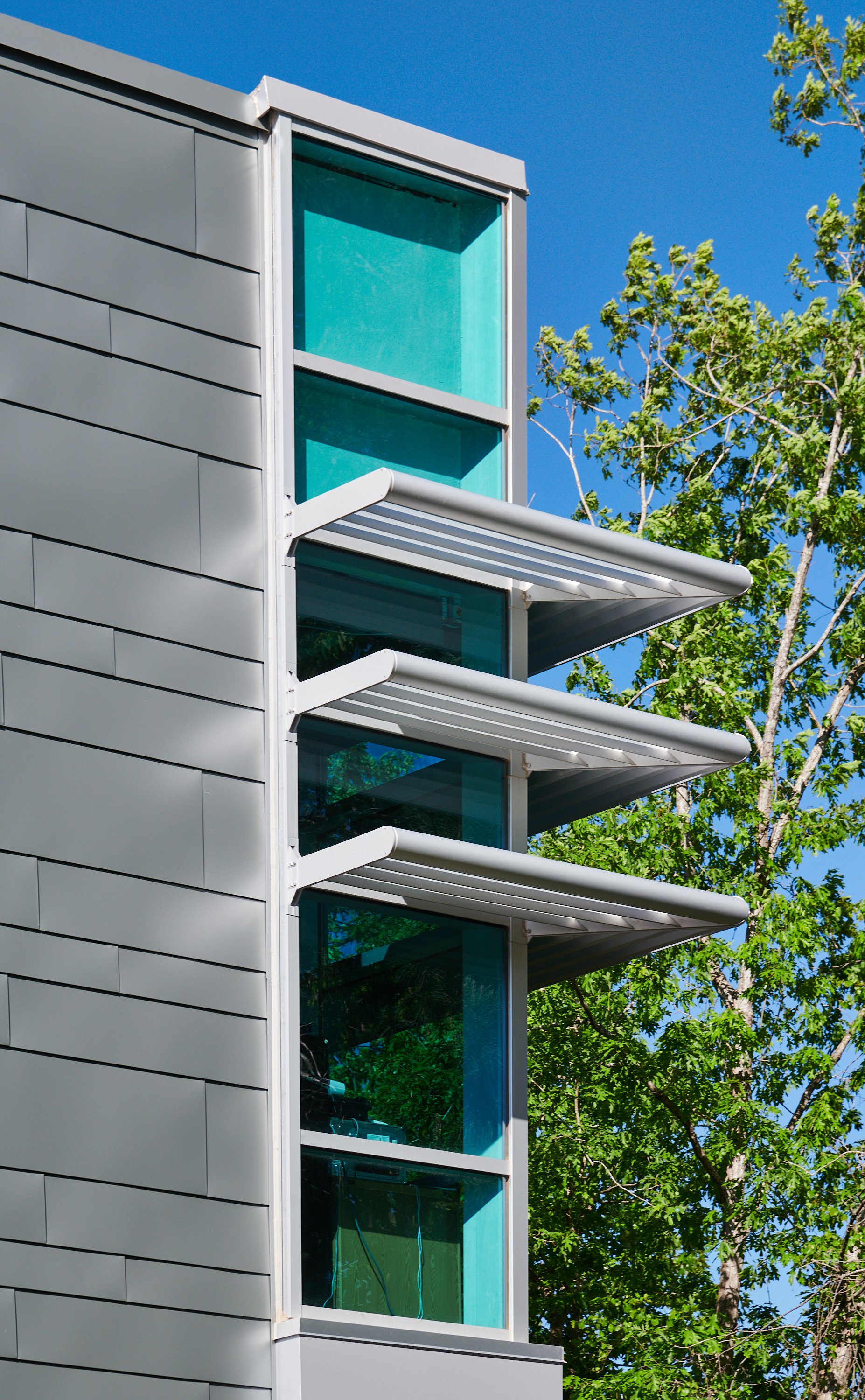Corner of a modern high school with varying sized lapped and seamed metal siding, curtainwall glass and sunshades, elevated over a street below - project by B Group Architecture, Inc.