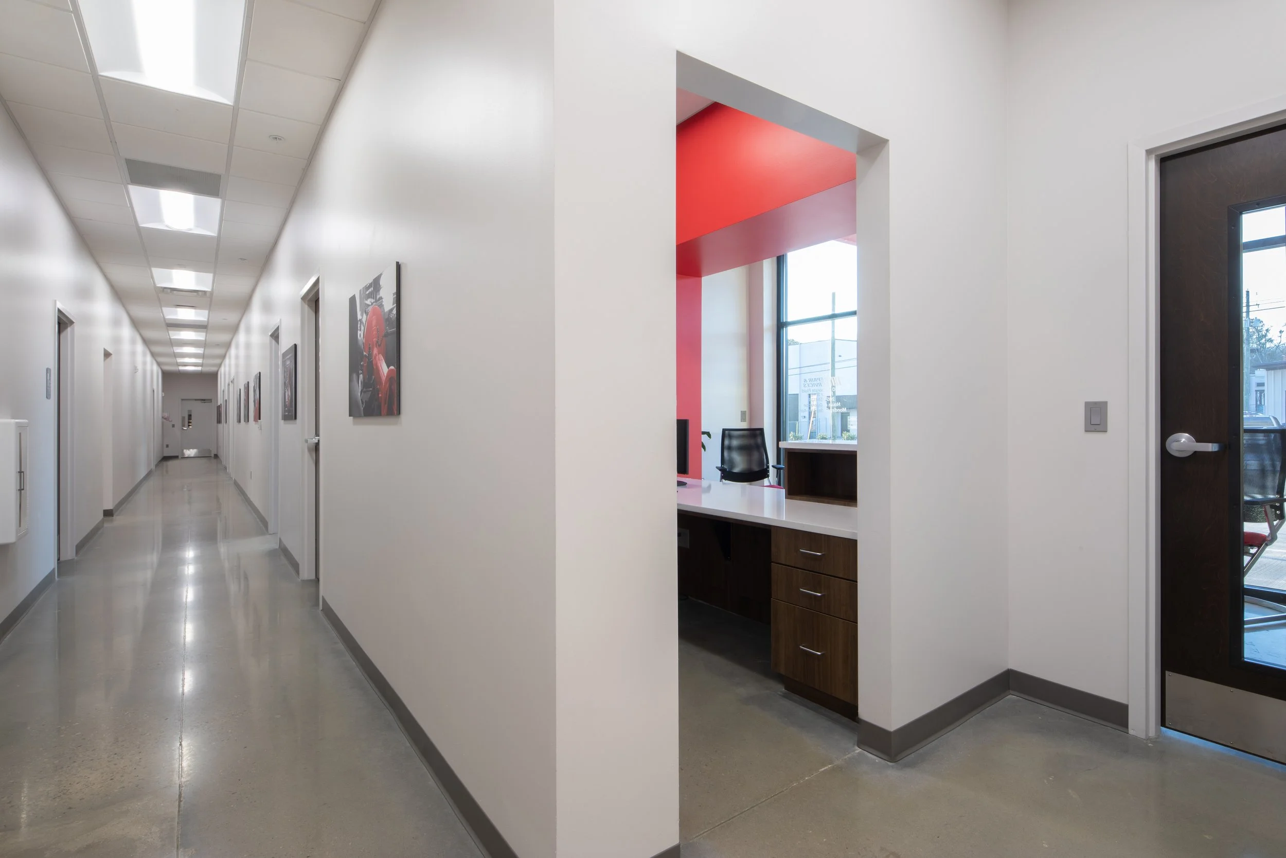 A clean, brightly lit office corridor with white walls, art on the walls, and a side office with pink interior walls, desk, and chair.