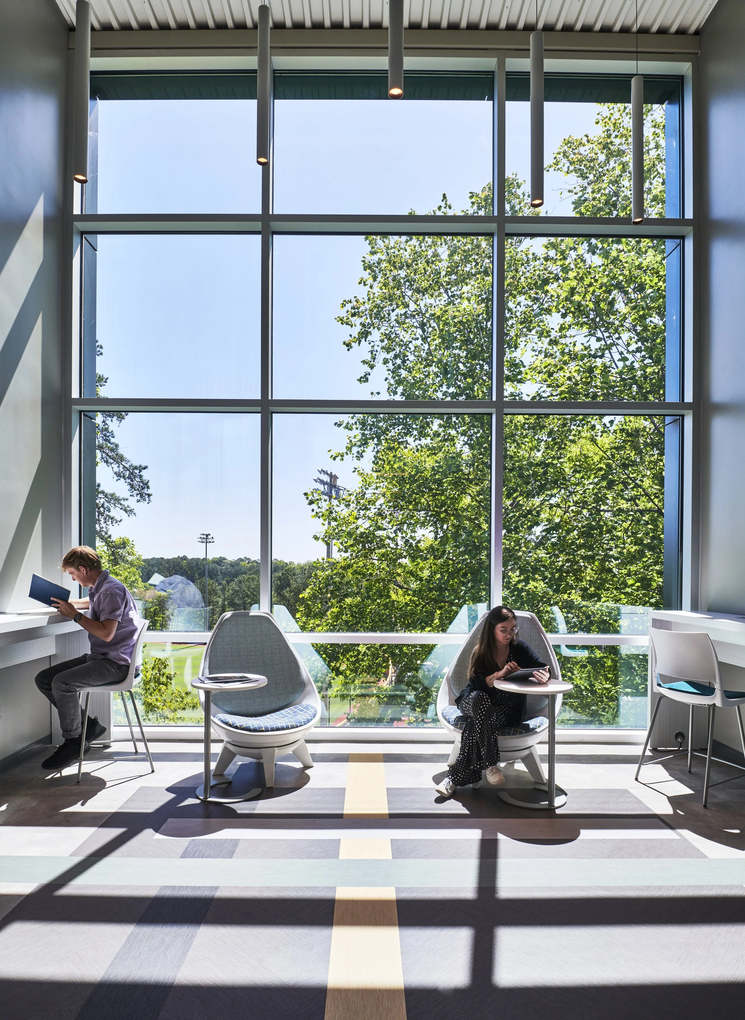 Interior of a modern lounge or study area with large window showing green trees and a clear blue sky, featuring two young people seated and reading or using devices.