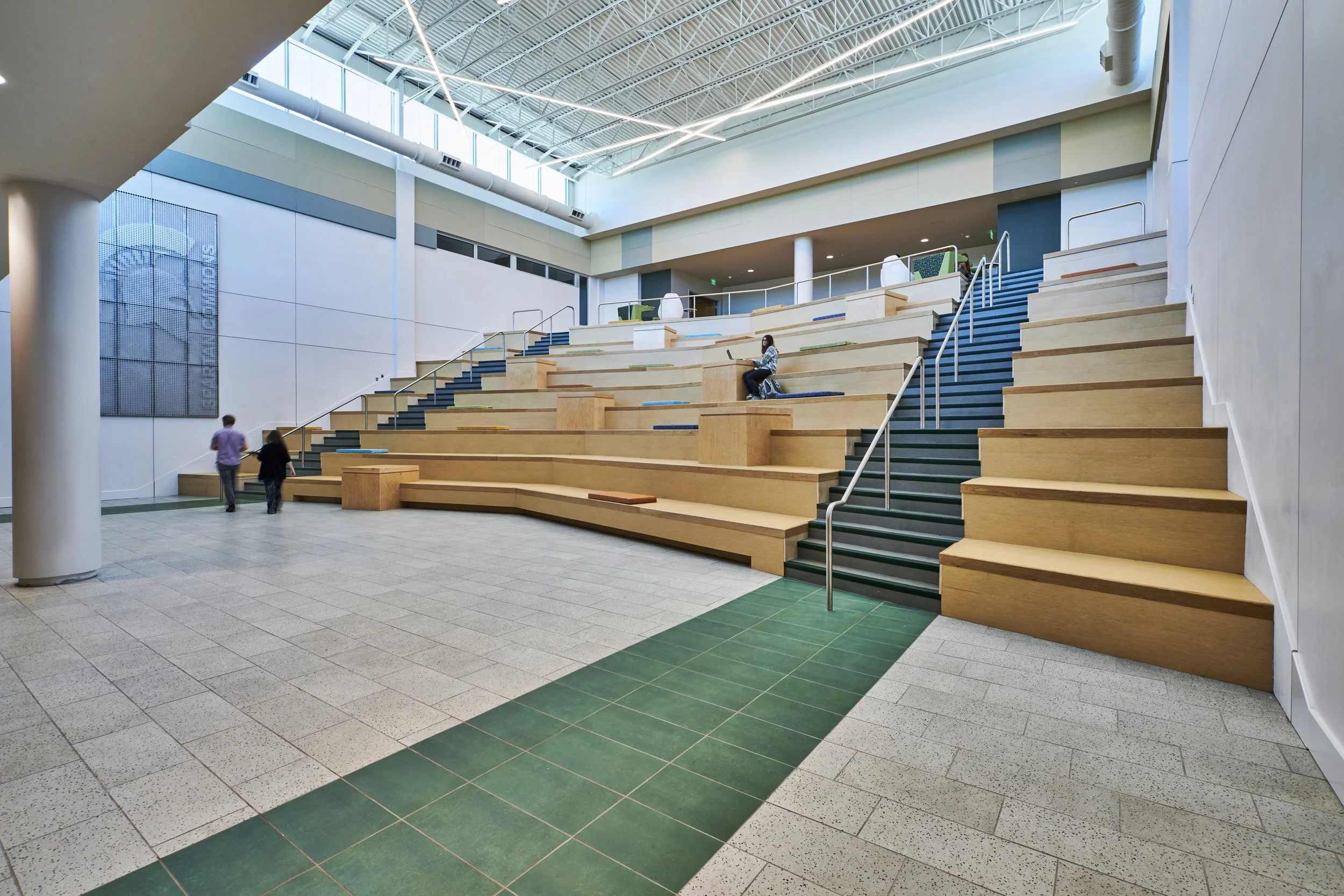 Interior view of a modern High School with the Spartan Commons tiered seating leading to a second level, showcasing large clerestory insulated windows- project by B Group Architecture, Inc.
