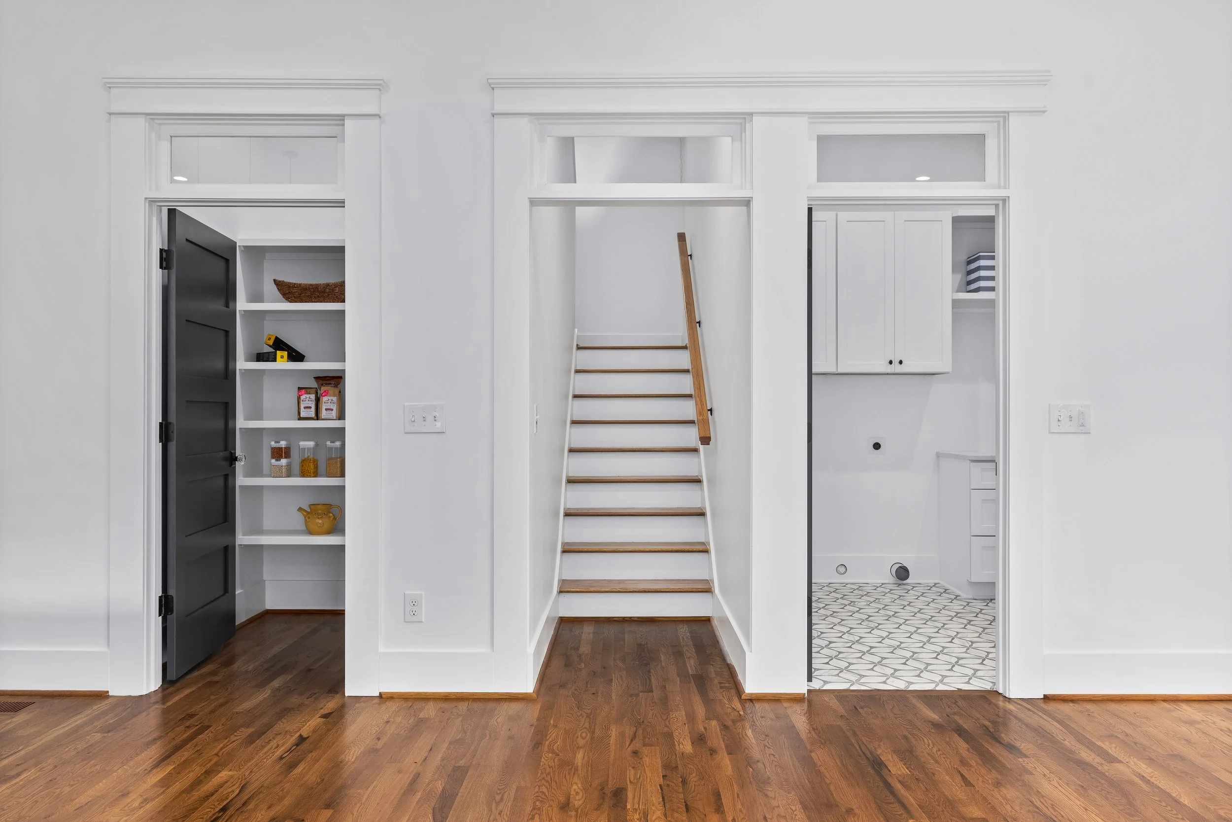 Interior view of a white-walled house showing a pantry, staircase, and laundry area, with hardwood flooring.