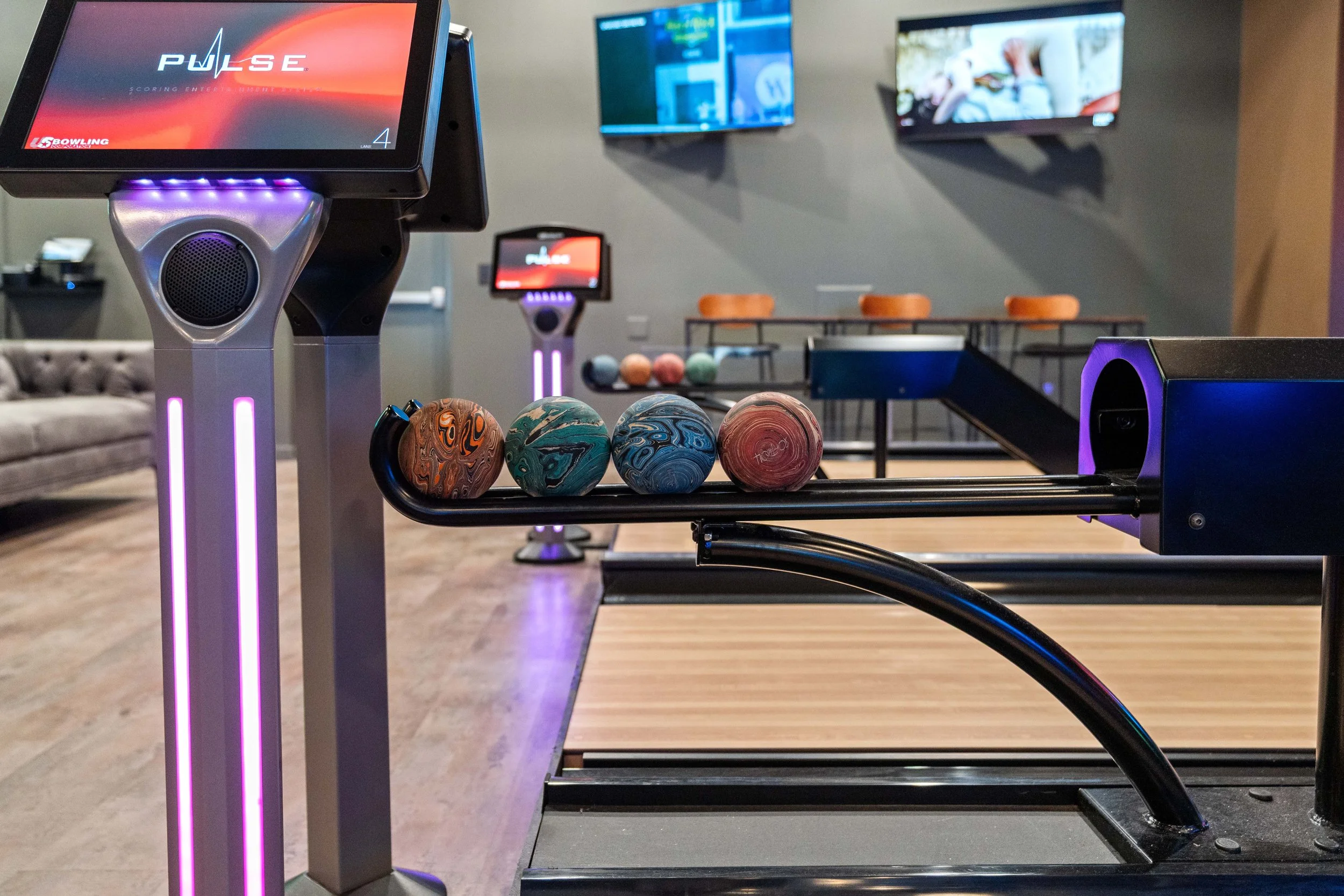 Duck Pin bowling balls on a ball return system, electronic scoring screens, and a seating area in the background. project by B Group Architecture, Inc
