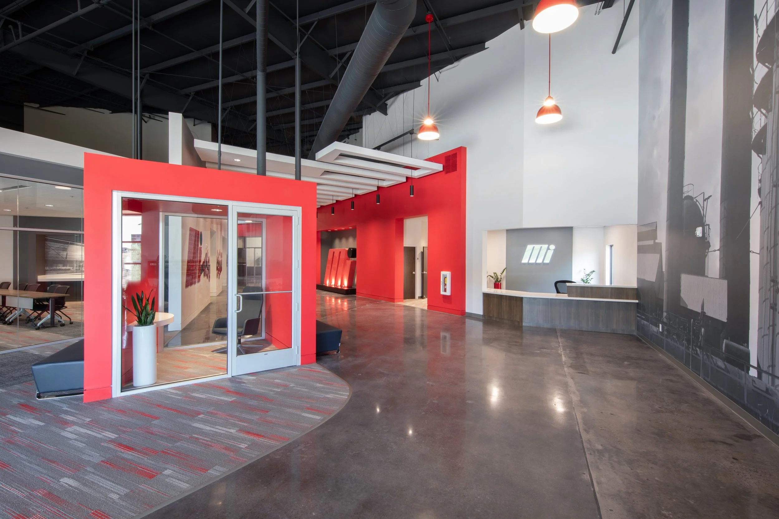 Modern lobby area with check-in desk, red accent wall, sitting area, glass entrance doors, and industrial ceiling with hanging lights. project by B Group Architecture, Inc. 