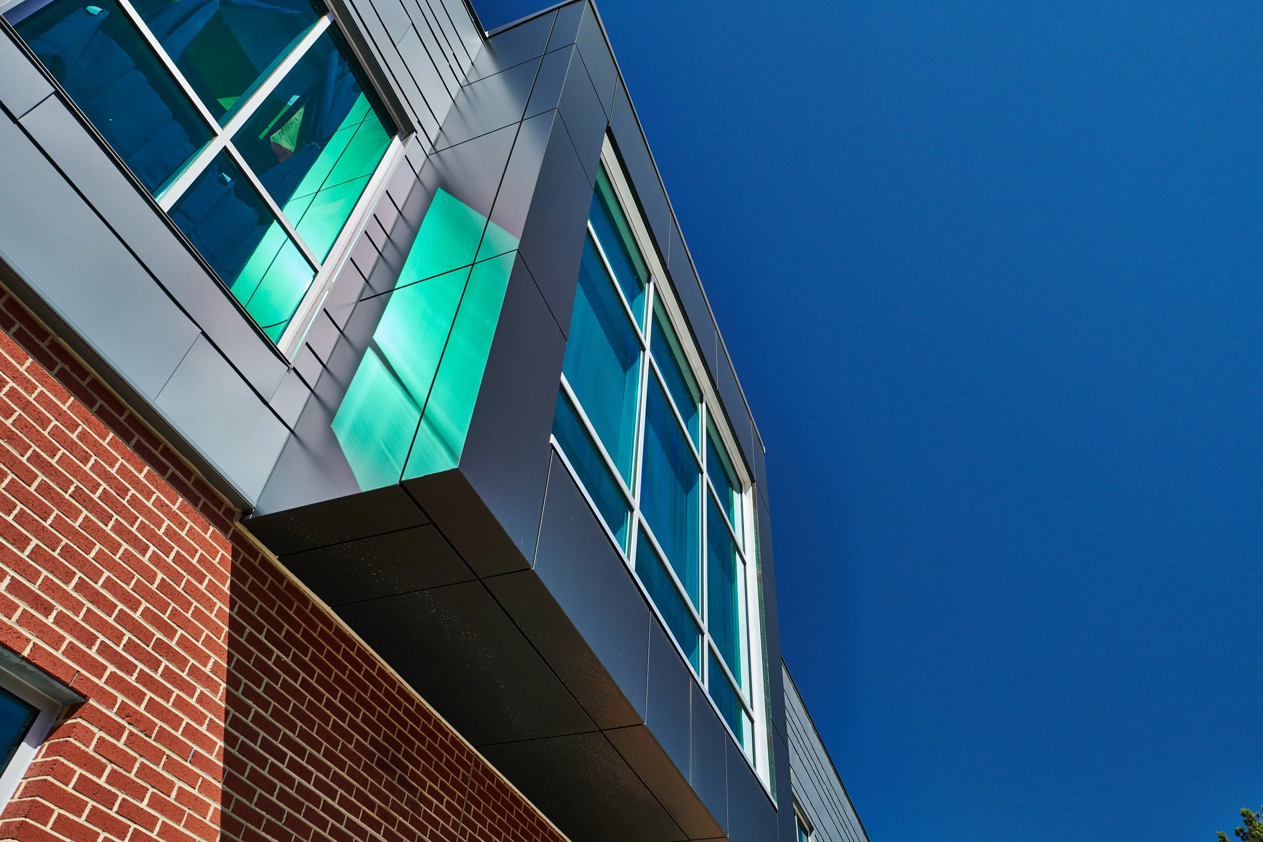 Close-up of a modern high school with brick lower walls, prefinished composite metal siding, and large glass windows against a clear blue sky.