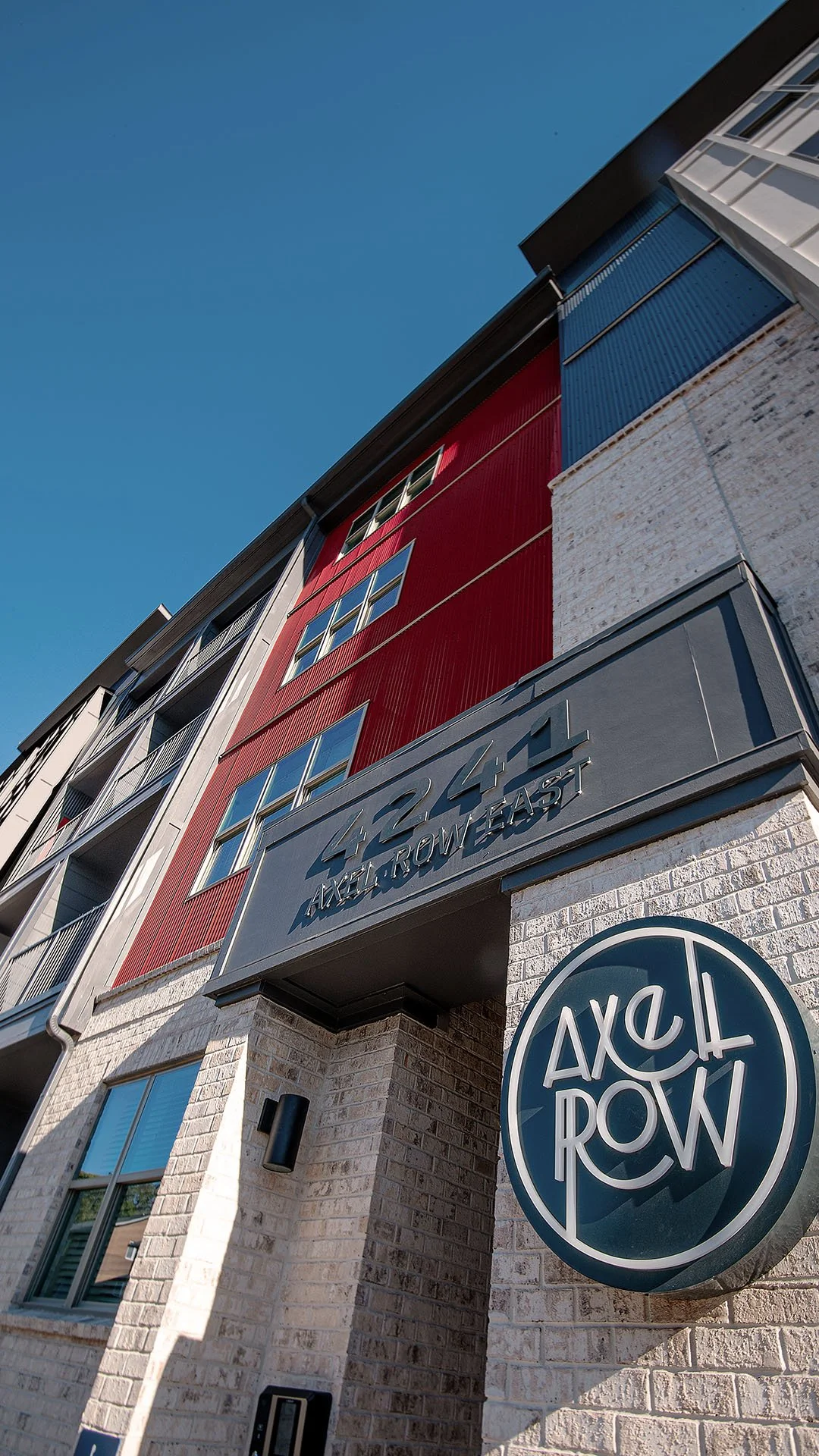 Low-angle view of a modern apartment building with red and  exterior siding, and stone showing the address 4241 and the name Axe & Row on a sign and round logo.  - project by B Group Architecture, Inc.
