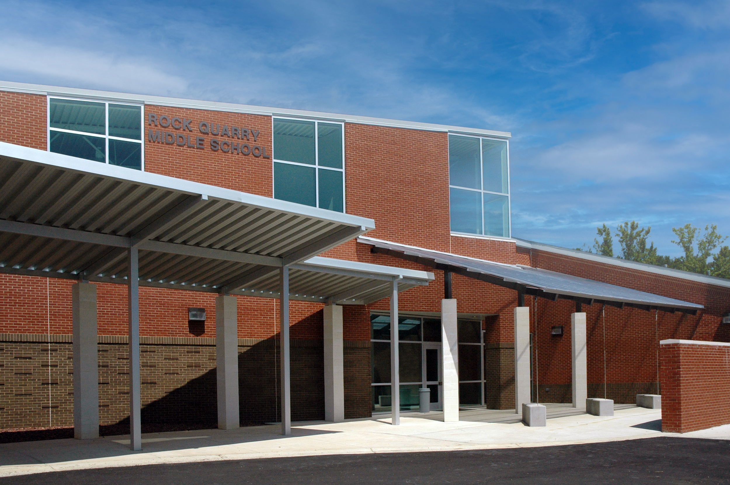 Front view of Rock Quarry Middle School building with brick walls, large windows, and a metal awning over the entrance, under a blue sky with some clouds.