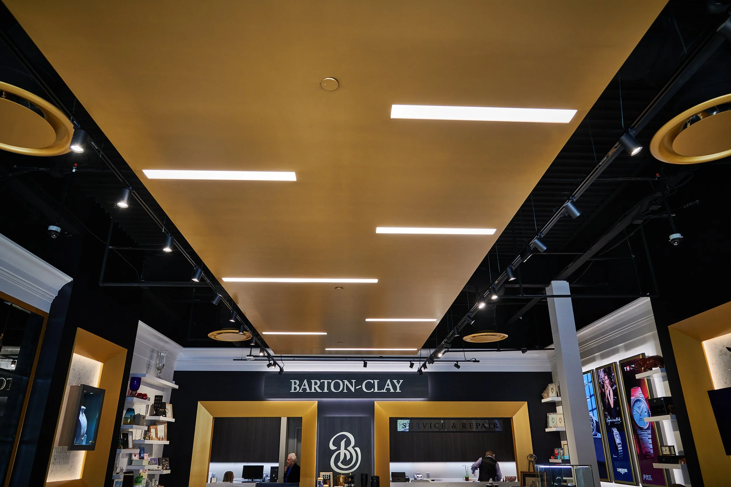 Interior of Barton-Clay with a black and gold color scheme, shelves and digital screens on the walls, and a floating gold ceiling with linear lights. project by B Group Architecture, Inc 