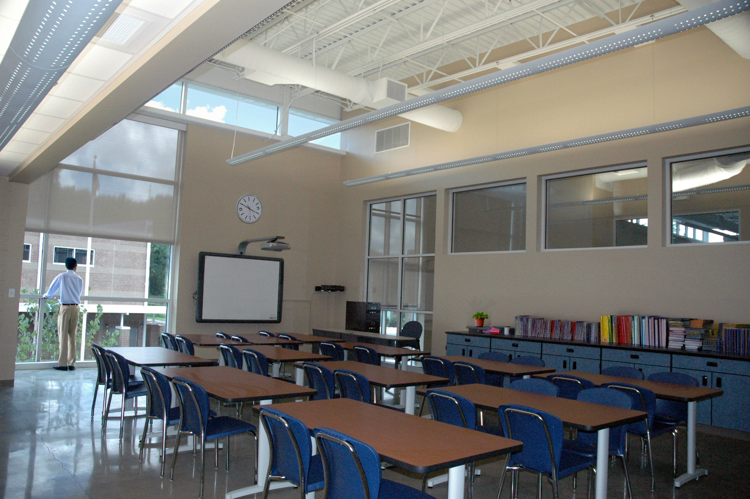 Classroom with blue chairs and brown tables, large windows, and a whiteboard. Open to structure above and clerestories accentuate the height of the space. - project by B Group Architecture, Inc.
