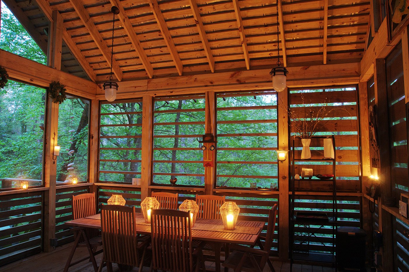 A cozy wooden screened-in porch for the Creekside Pavilion with slat wall design located in a lush green forest, illuminated from inside. project by B Group Architecture, Inc.
