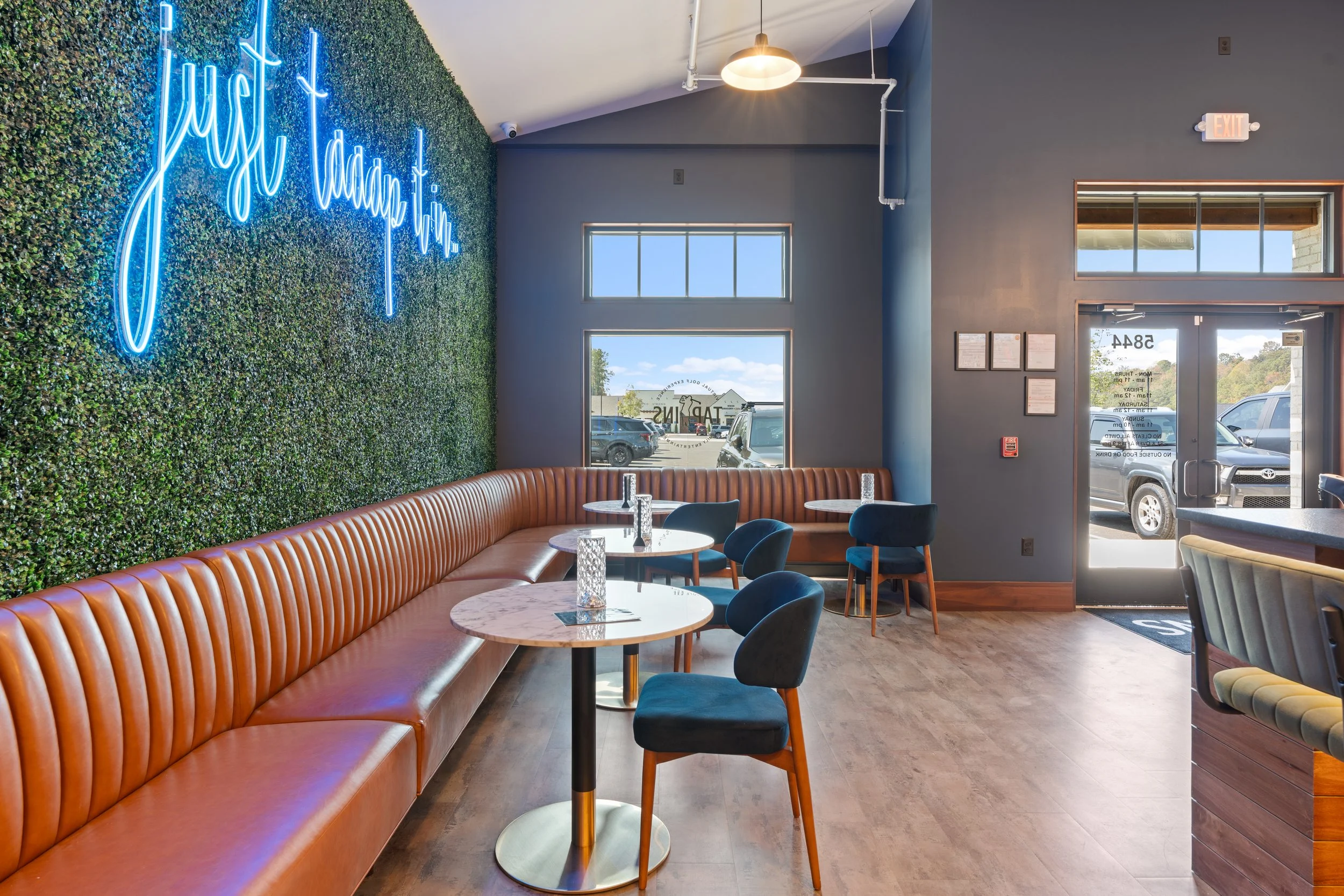 Interior of a modern lounge and sports  bar with blue walls, a green plant wall with neon sign, brown leather bench seats, and small round tables with blue  velvet chairs. project by B Group Architecture, Inc

