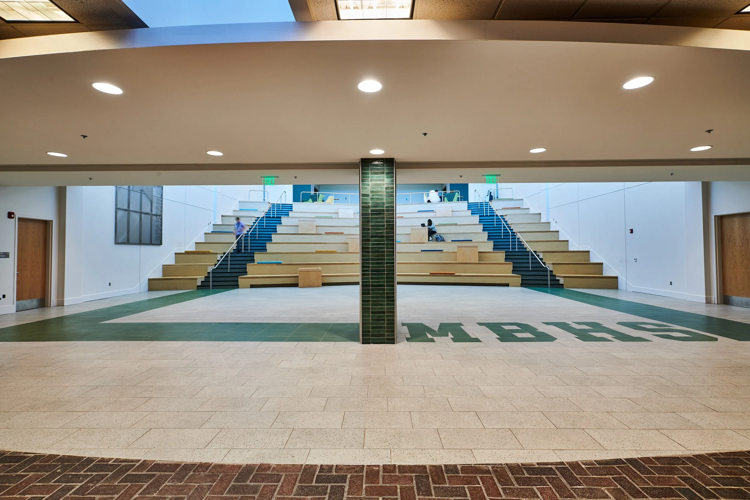 Interior view of a modern High School with the Spartan Commons tiered seating leading to a second level, featuring a custom school colored accent tile design - project by B Group Architecture, Inc.