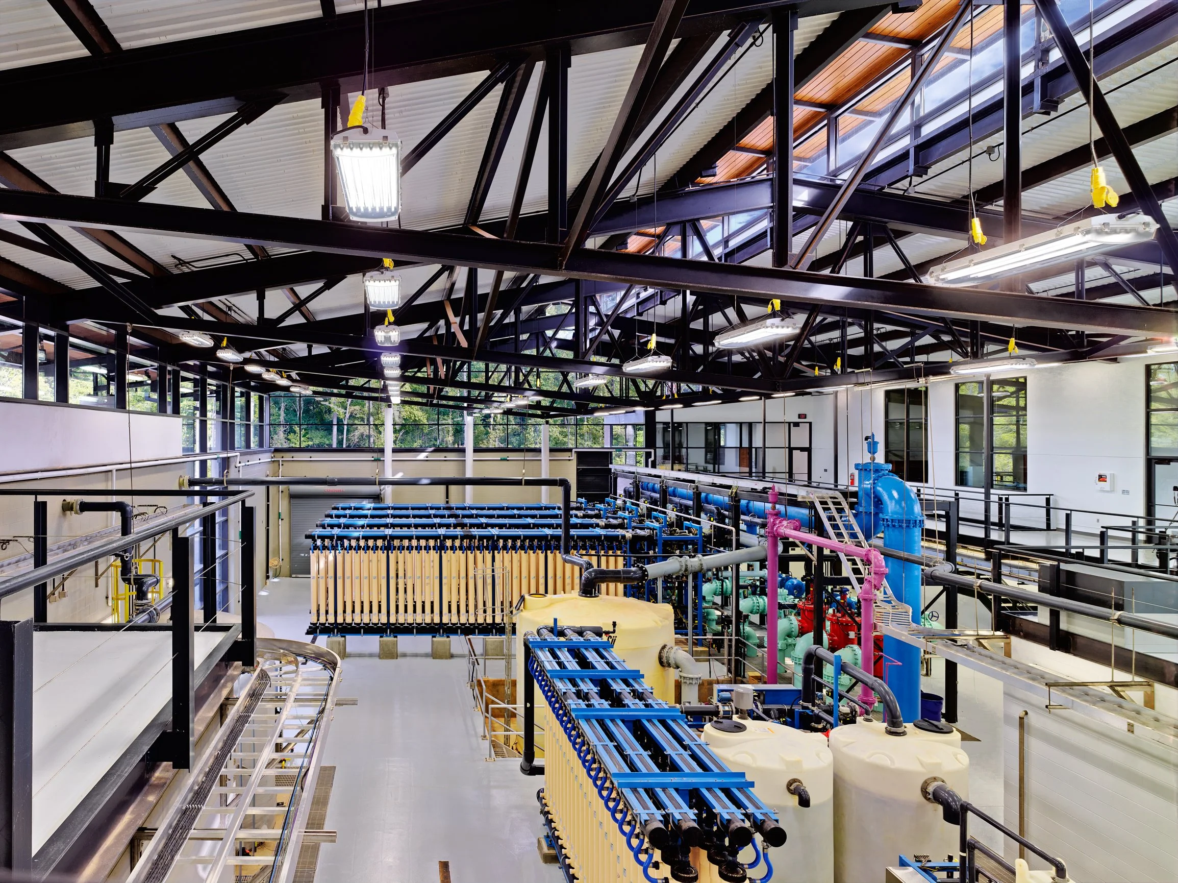 Industrial water treatment facility with pipes, tanks, and filtration equipment inside a spacious building with large windows and a metal roof structure. project by B Group Architecture,  Inc.