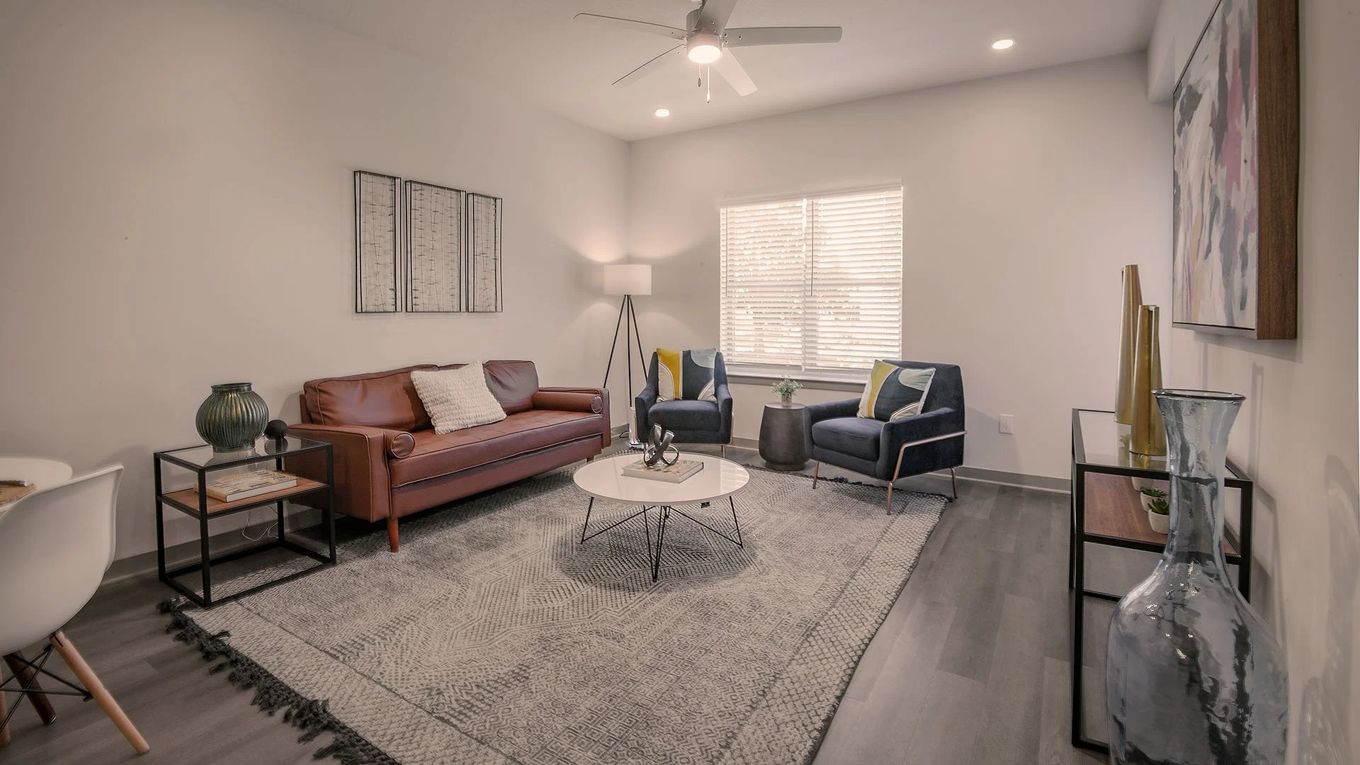 Modern living room  in Axel Row Apartments with a brown leather sofa, two dark blue armchairs with dark and yellow pillows, a white round coffee table with decorative items, a floor lamp, a window with wooden blinds, wall art, a large glass vase, and