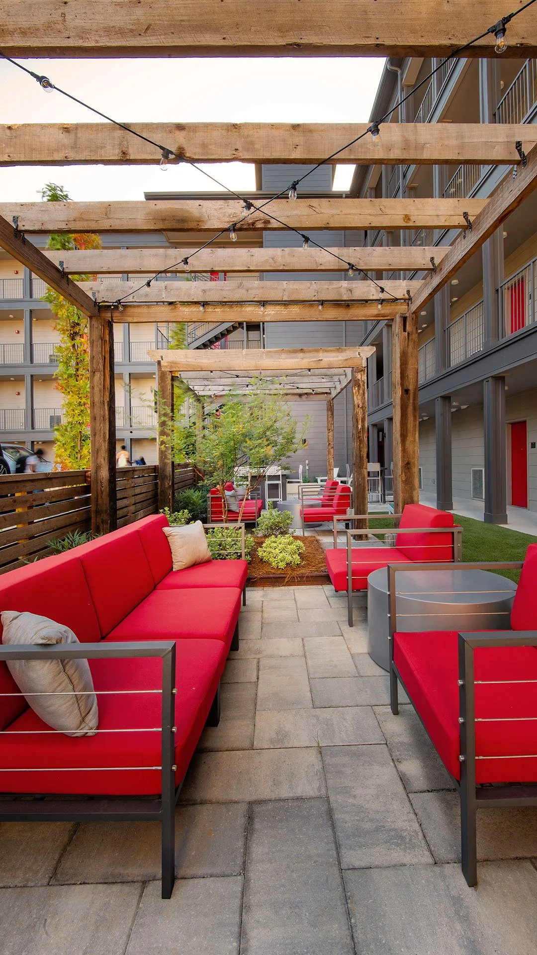 Outdoor patio area with red cushioned seating, potted plants, and wooden pergola with string lights in a modern apartment complex. project by B Group Architecture, Inc.