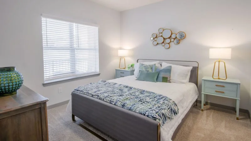 One bedroom in Axel Row Apartments with a gray bed, white bedding, and decorative pillows. Two blue nightstands with lamps are on each side of the bed. There is a gold circular wall art above the bed, a window with white blinds, and a wooden dresser 