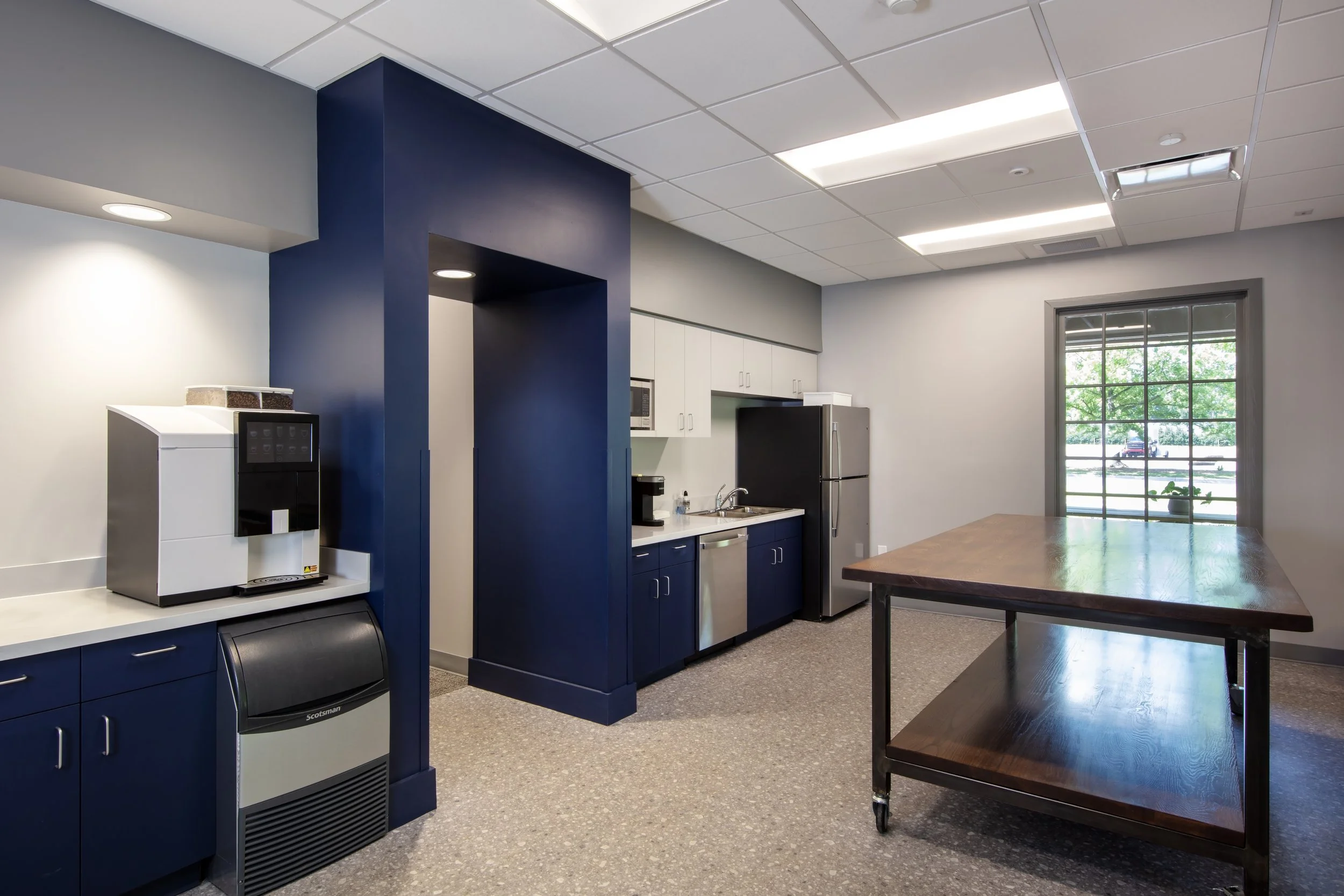 Kitchen area with blue cabinets, white upper cabinets, a stainless steel refrigerator, black coffee maker, coffee machine, and a large wooden table with wheels.