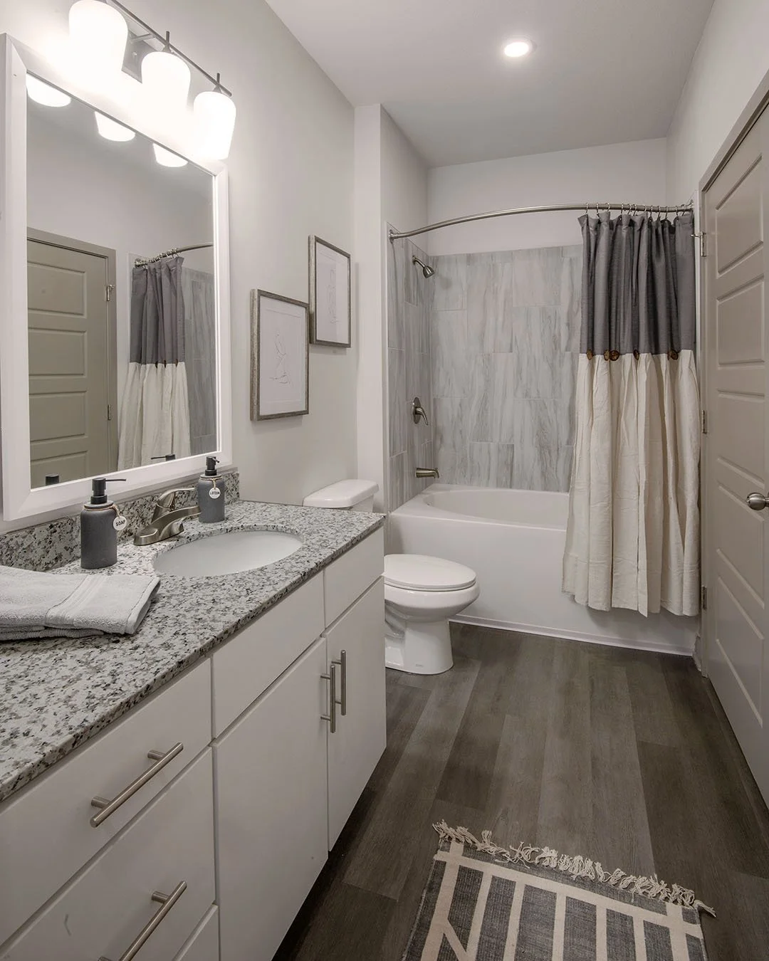 Modern bathroom with a granite countertop vanity, large mirror, framed artwork, and a combination bathtub and shower with gray and white tiles, gray shower curtain, and dark wood flooring. - project by B Group Architecture, Inc.