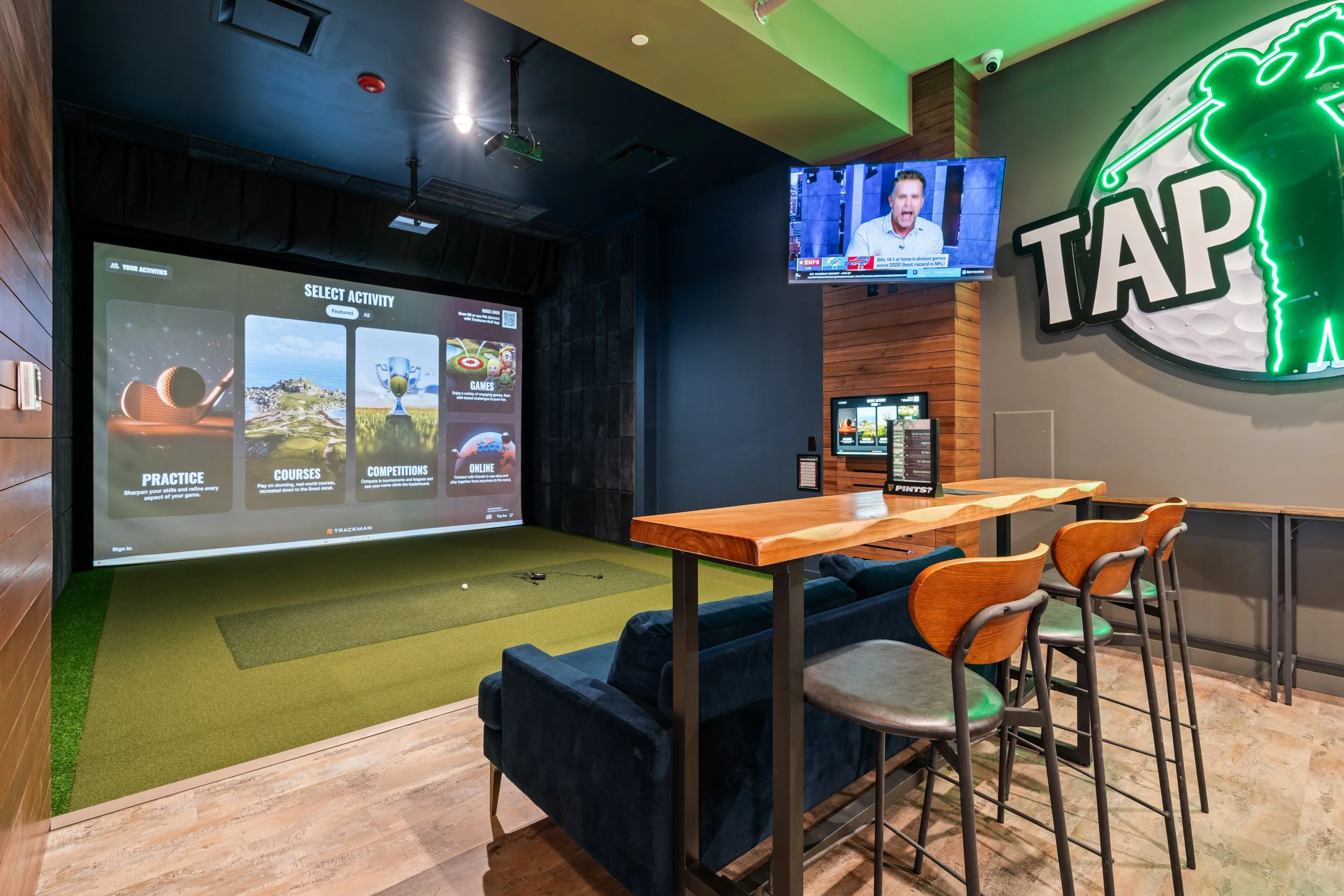 Indoor golf simulator sport's bar with a large screen display for practice, courses, competitions, and online games. Wooden bar-height chairs and a sofa are positioned near a counter. There is a television mounted on the wall showing a man speaking. 