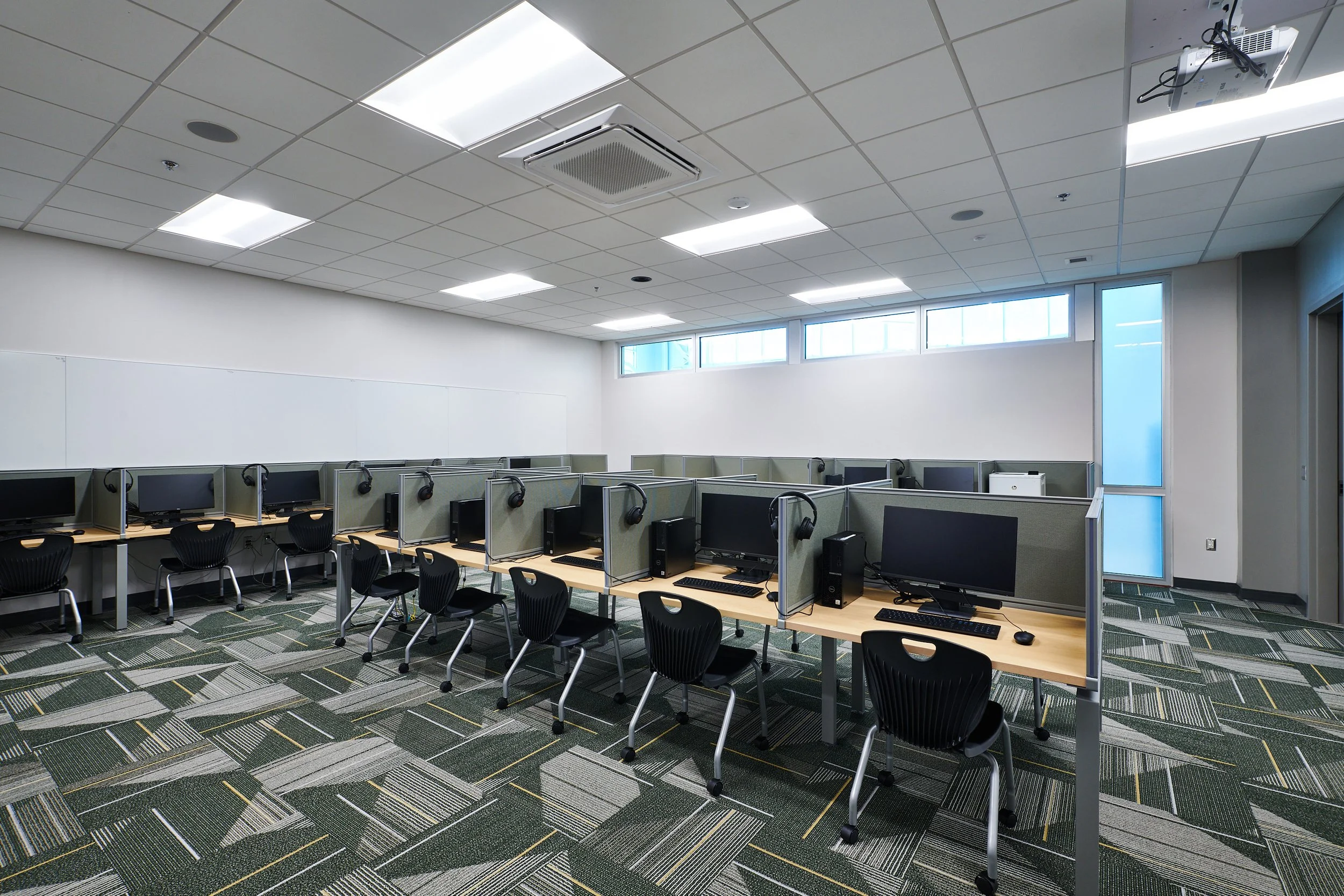 Computer lab with multiple workstations, each with a monitor, keyboard, mouse, and headphones, arranged in rows with cubicle dividers, and a windowed wall in the background. Custom carpet tile to match Mountain Brook High School colors. - project by 