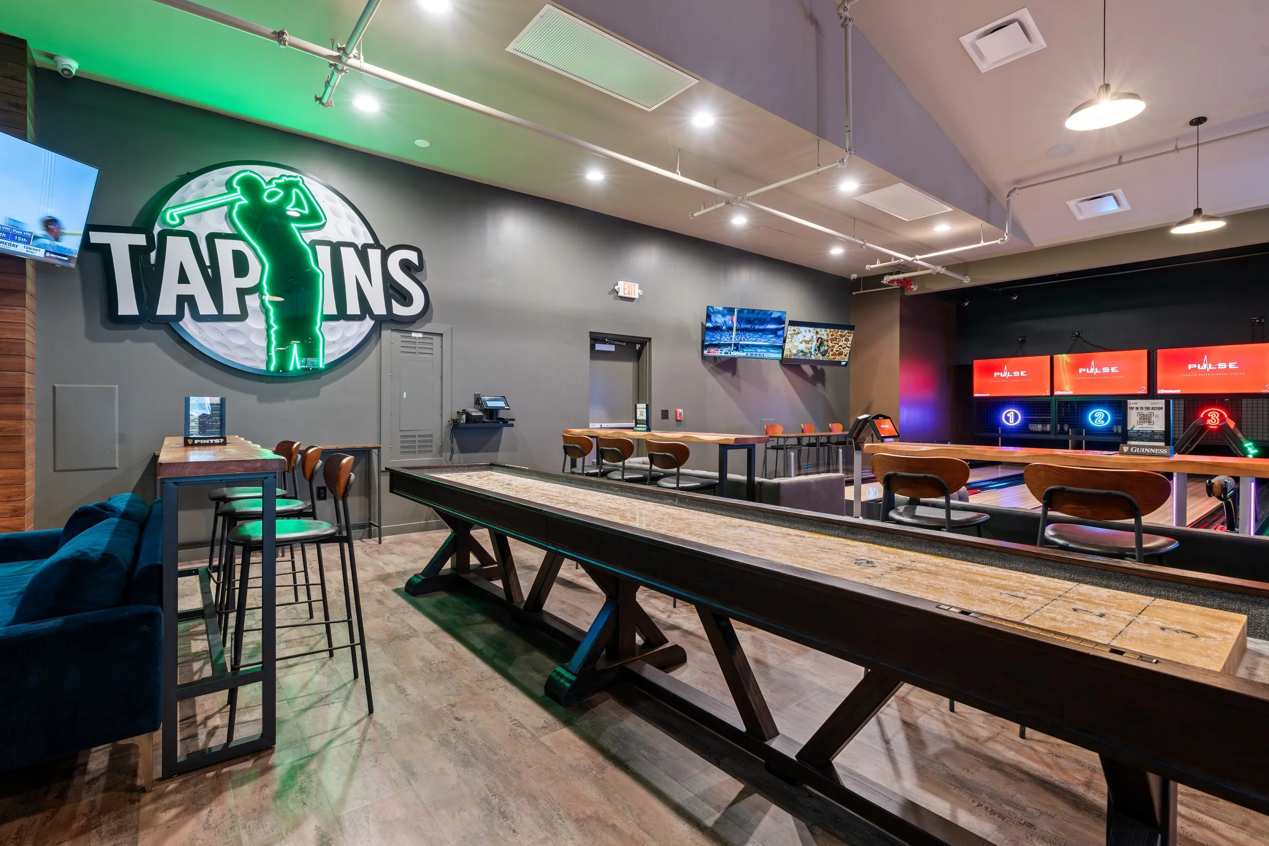 Indoor golf entertainment venue with a large illuminated sign reading "TAPINS". There are multiple viewing screens showing sports, a mechanical shuffleboard table in the foreground, and duck pin bowling lanes in the background with illuminated scorin