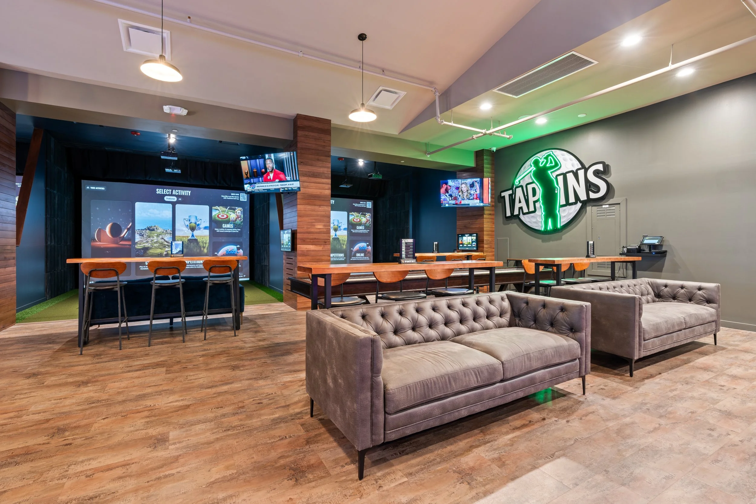 An indoor entertainment lounge with a large illuminated sign reading 'TAP INS' featuring a green glowing bowling pin figure. There are sofas, several bar-height tables with chairs, and multiple TV screens displaying games and sports. project by B Gro