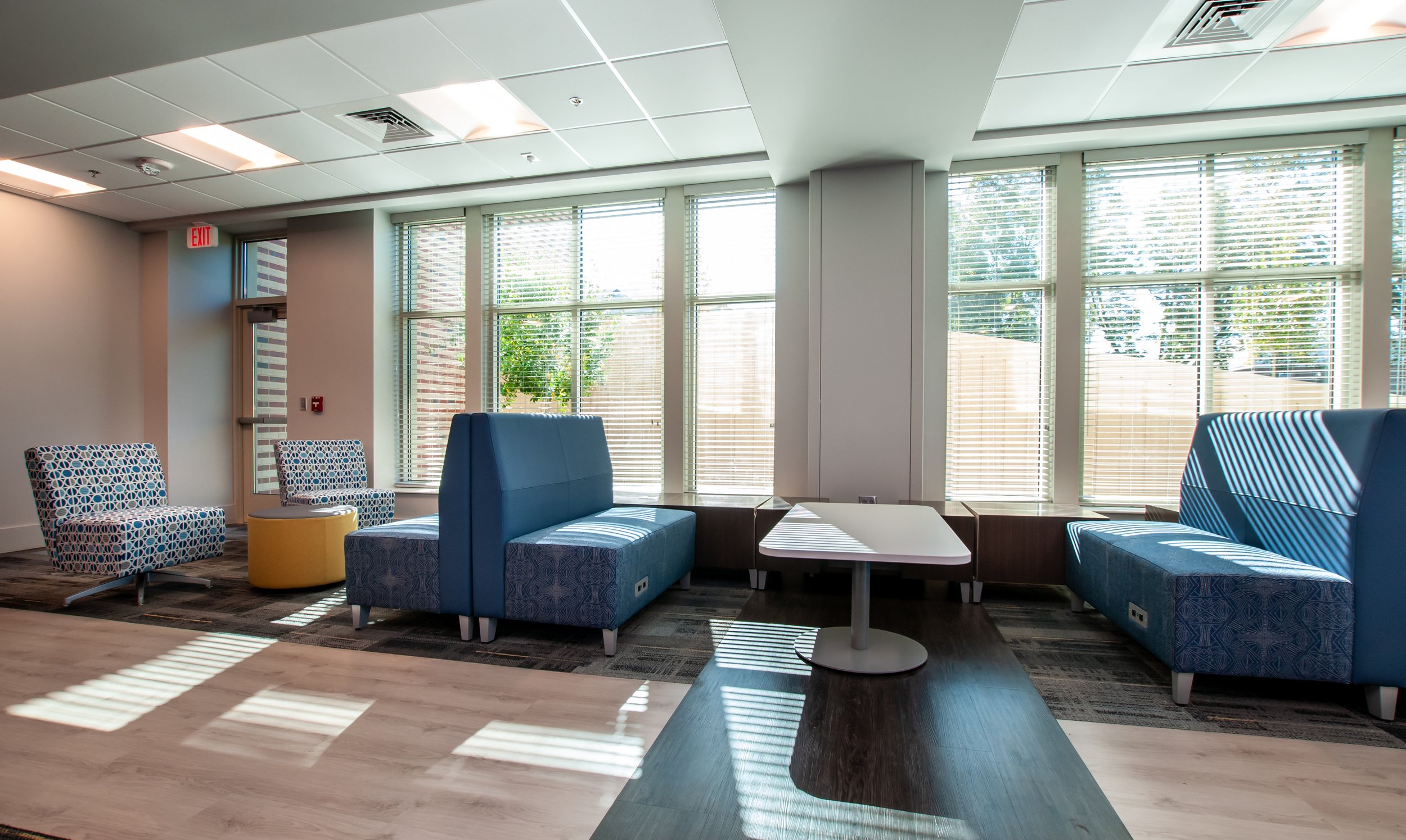 A modern lounge area with blue and patterned chairs, yellow ottoman, and a white table, illuminated by natural light from large windows with blinds.