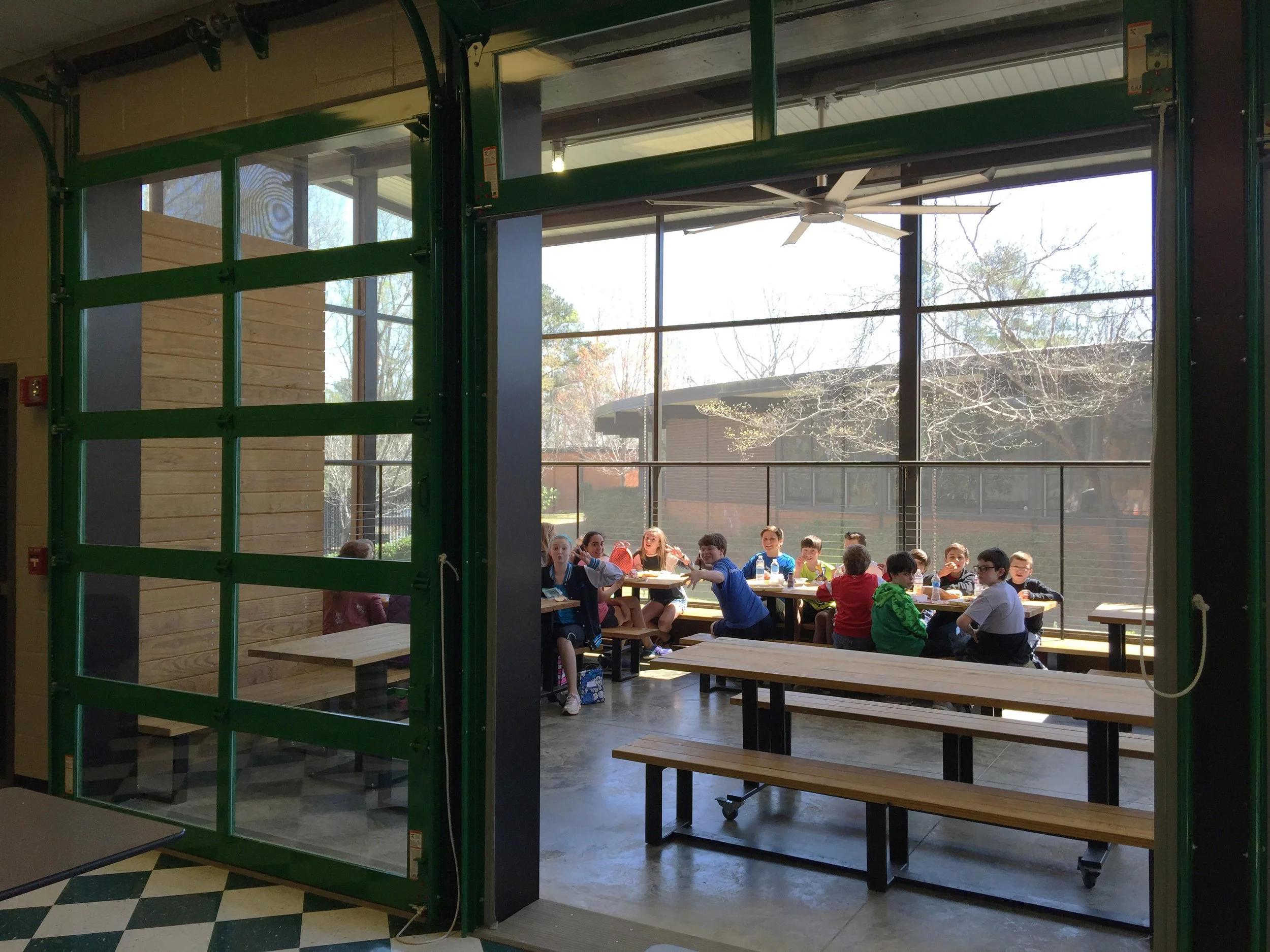 A modern glass dining porch at Brookwood Forest  Elementary School during sunset. Green roll-up doors and a large fan. project by B Group Architecture, Inc. 