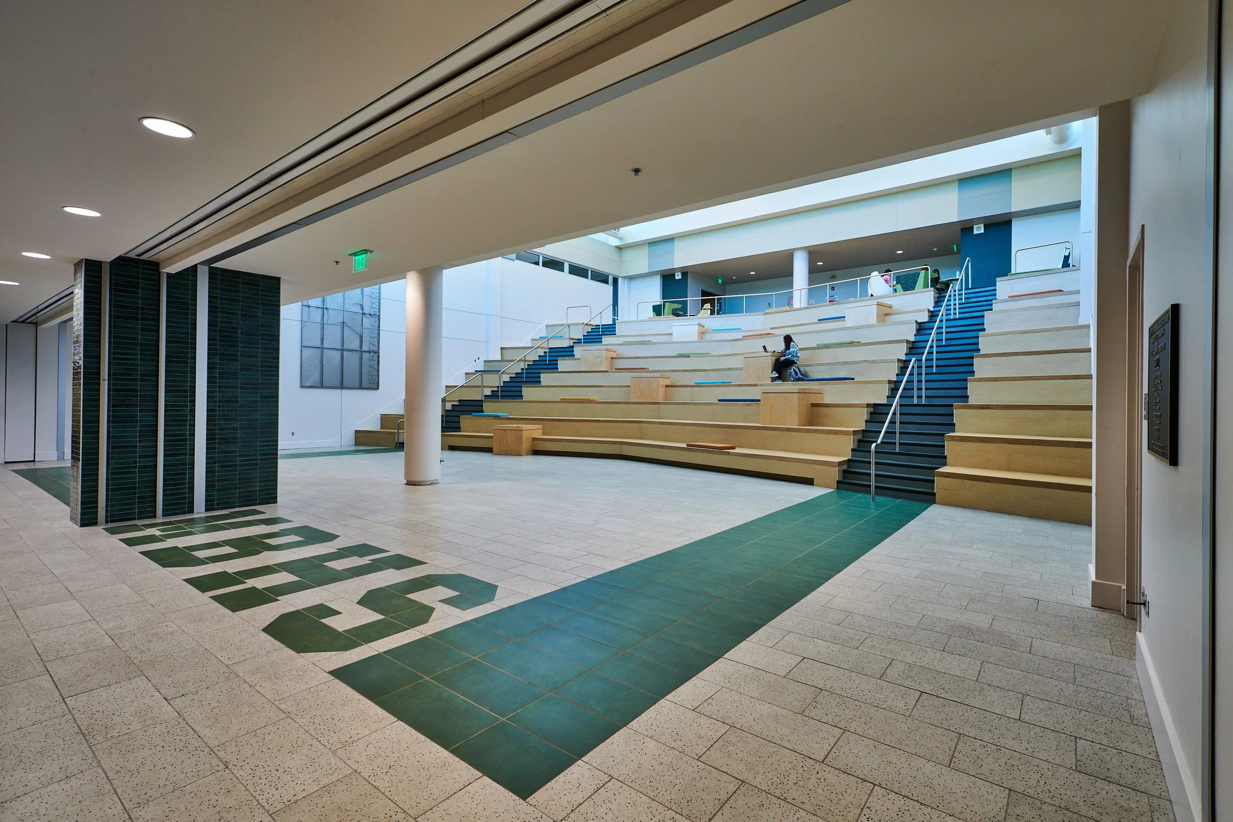 Interior view of a modern High School with the Spartan Commons tiered seating leading to a second level, featuring a custom school colored accent tile design - project by B Group Architecture, Inc.
