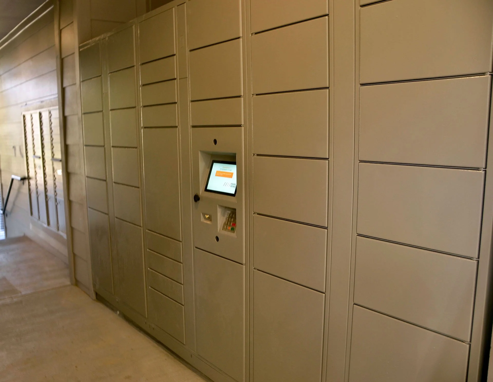 Self-service parcel locker with multiple compartments and a digital screen in a building corridor.