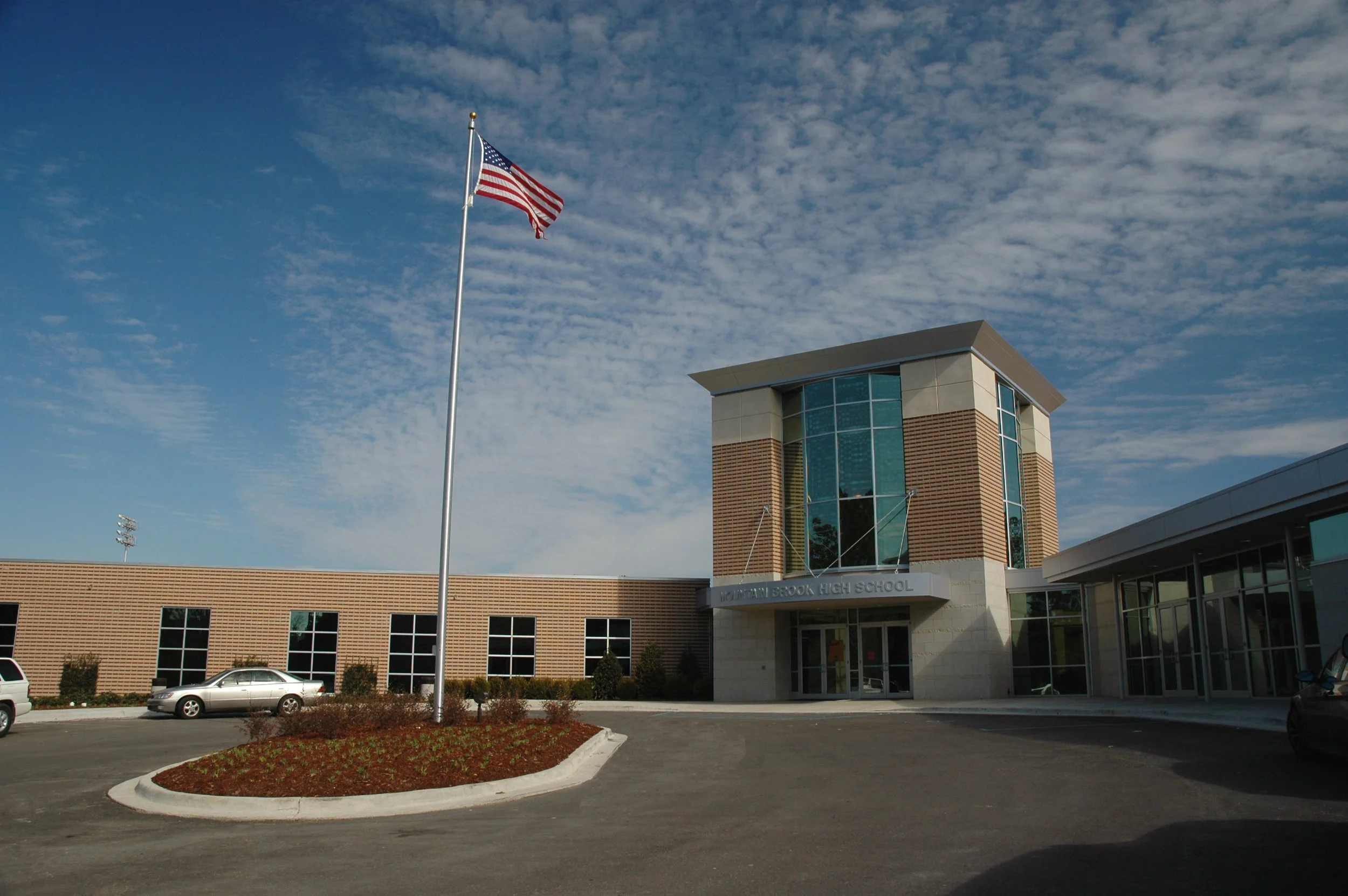 The exterior entrance of Mountain Brook High School with the American flag. The use of varying brick colors in rows is seen throughout campus. Cast stone stone and curtainwall frame the entrance.- project by B Group Architecture, Inc.