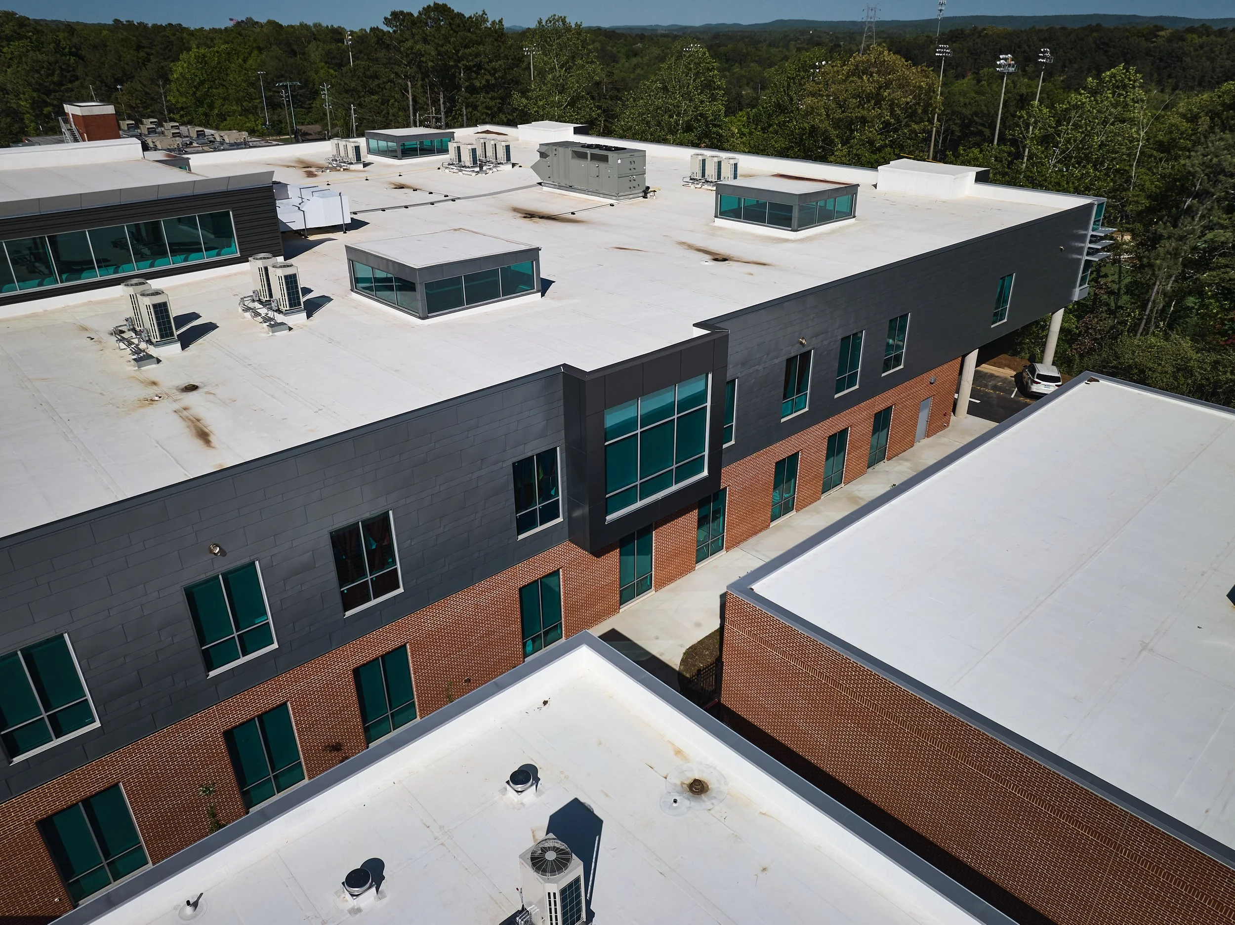 Aerial view of Mountain Brook High School with a flat white TPO roofs, multiple windows including clerestories, and HVAC units in a green area with trees and a sports field in the background - project by B Group Architecture, Inc.