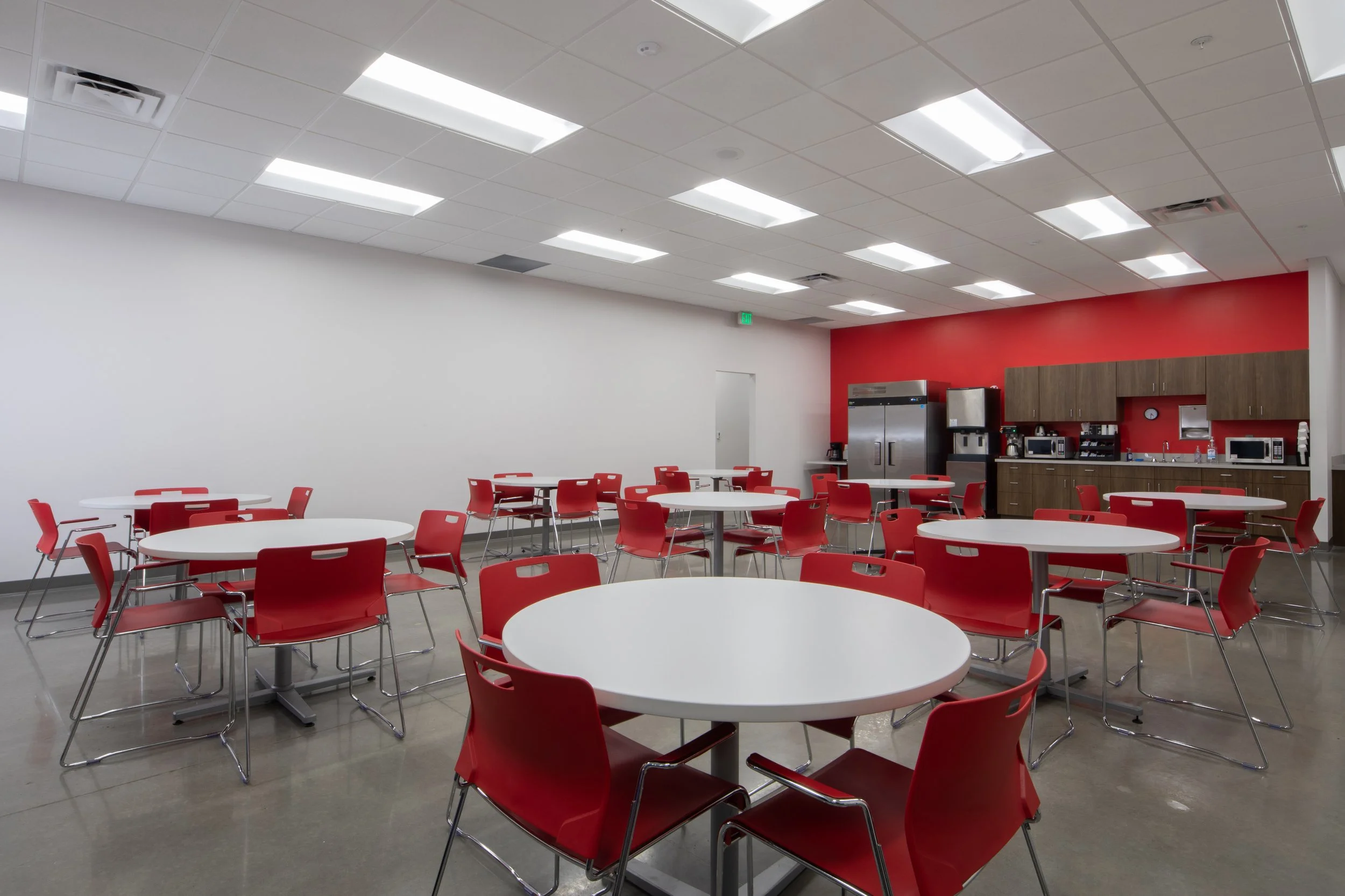 A break room at  Motion Industries  Mi logo on the wall. project by B Group Architecture, Inc.. with round white tables and red chairs, a kitchen area with brown cabinets, and a red accent wall. project by B Group Architecture, Inc..