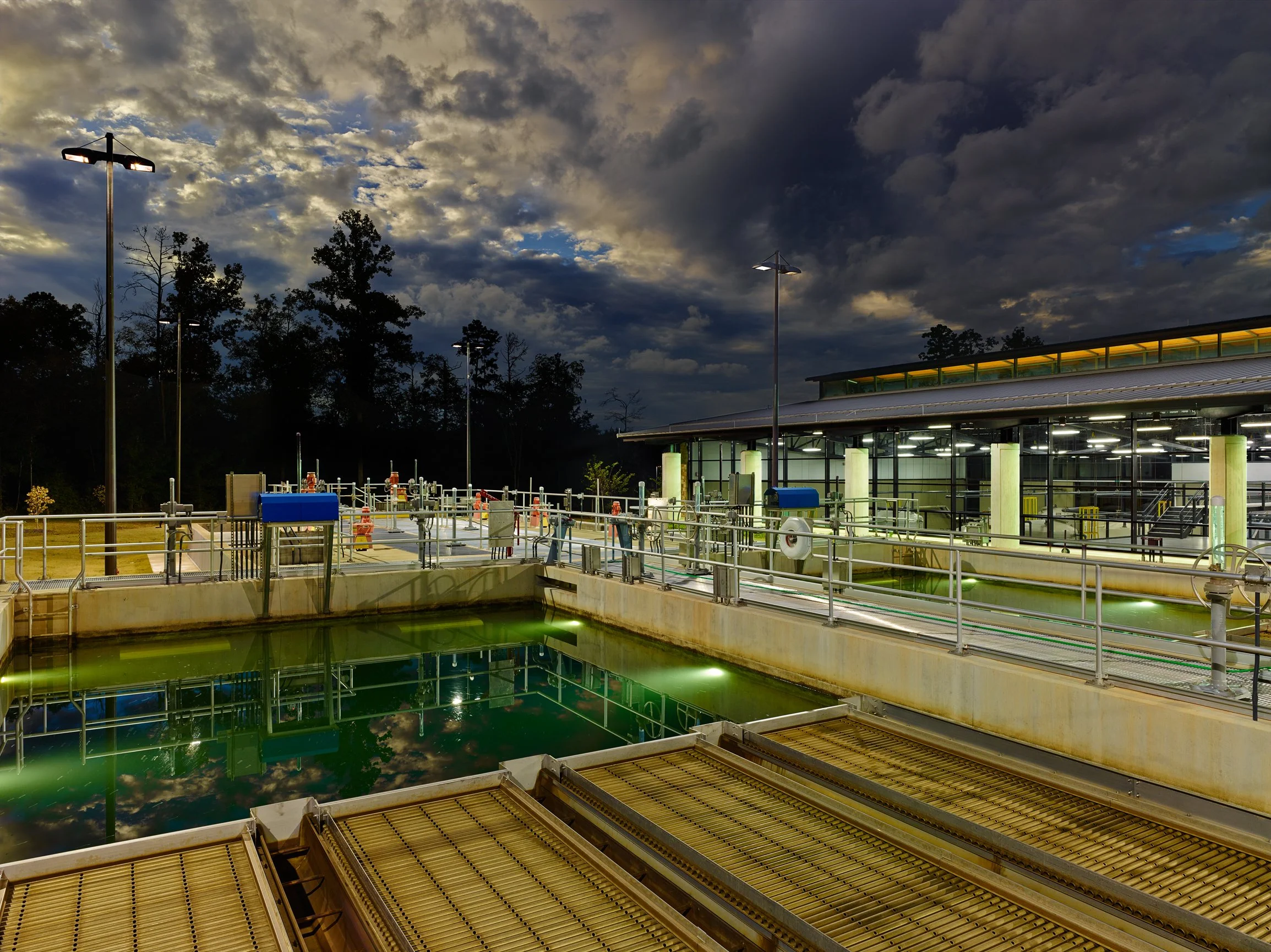 A wastewater treatment plant with illuminated tanks, pipes, and a building, under a cloudy sky. -project by B Group Architecture,  Inc.