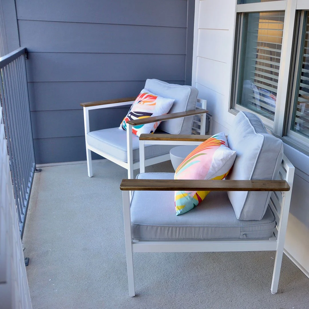 Two outdoor chairs with colorful pillows on a balcony with gray walls, beige carpet, and a window with blinds. - project by B Group Architecture, Inc.