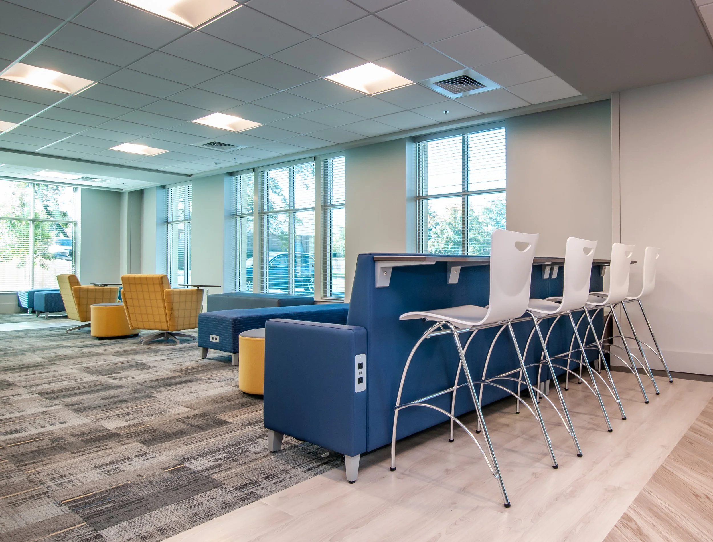 Modern lounge area with colorful armchairs, bar-height chairs along a counter, large windows with blinds, and patterned carpet flooring.
