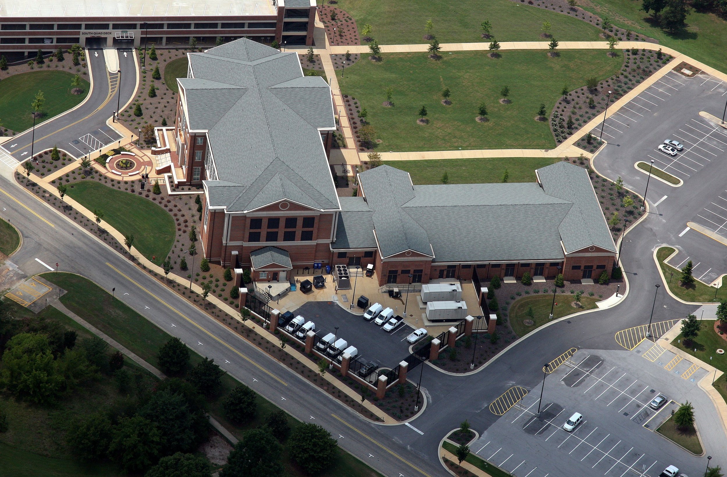 Aerial view of a university building with a pitched metal roof construction, surrounded by parking lots and green landscaped areas with trees and walkways.- project by B Group Architecture, Inc.
