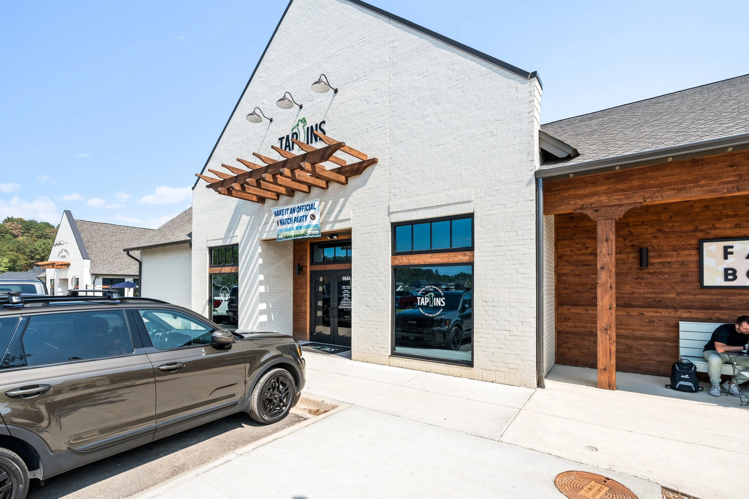 Exterior of Tap Ins Tap Ins sports bars with a white brick facade, large window with the Tap Ins logo, a wooden pergola above. project by B Group Architecture, Inc.