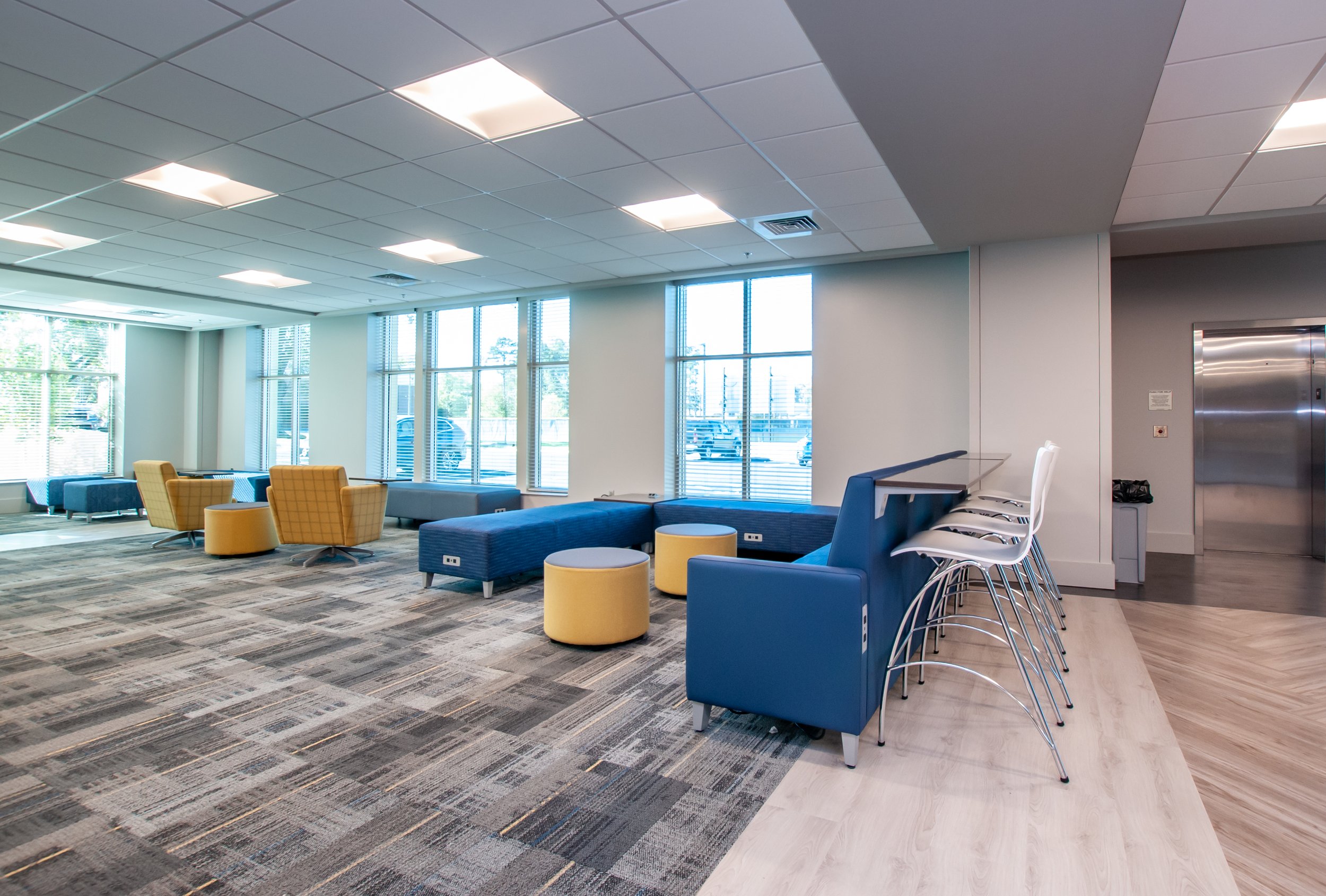 Modern waiting area with colorful chairs, ottomans, and a bar-height counter with white bar stools by large windows in a bright library space.