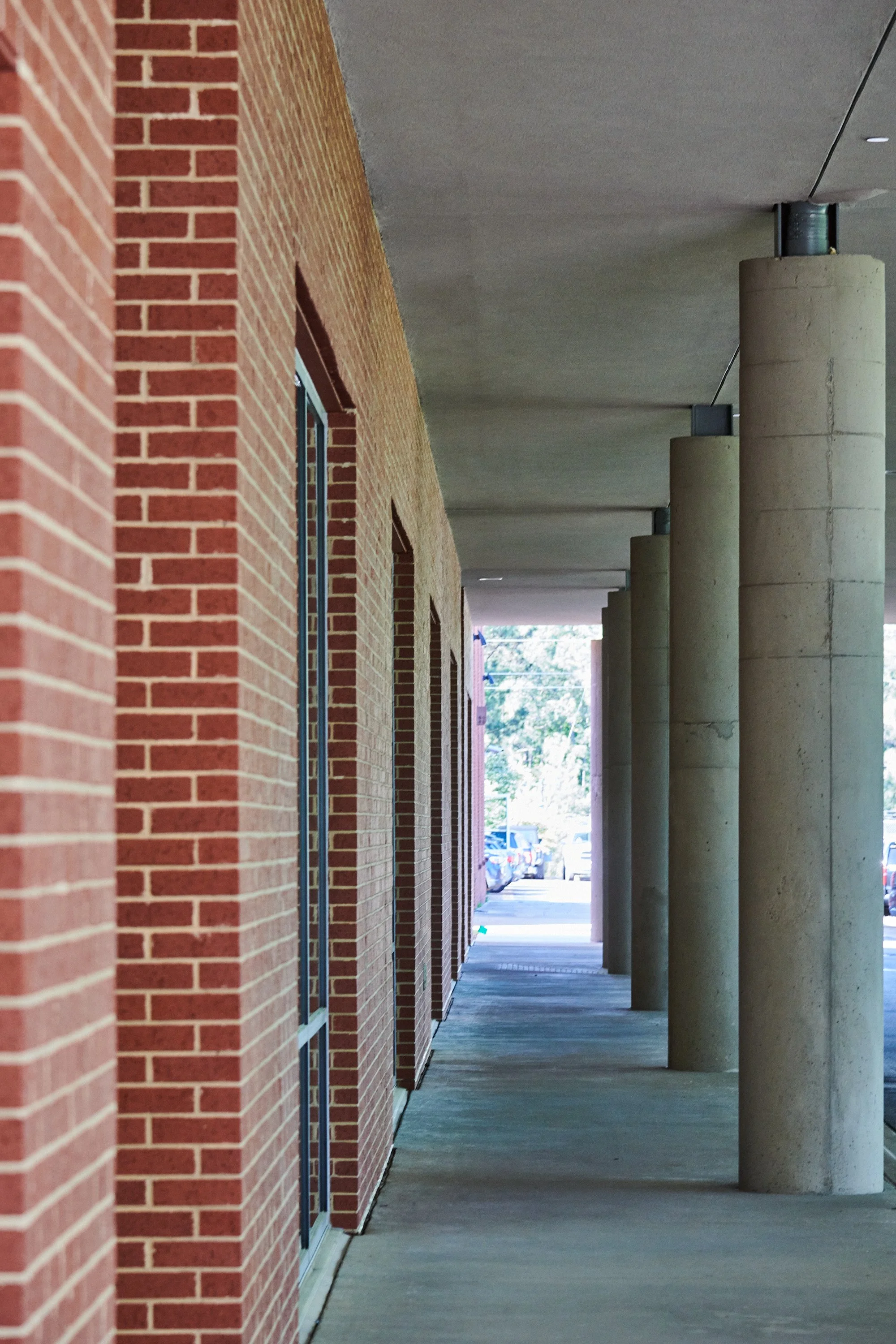 A sidewalk with brick walls on the left and concrete pillars on the right, leading toward a parking lot in the background- project by B Group Architecture, Inc.