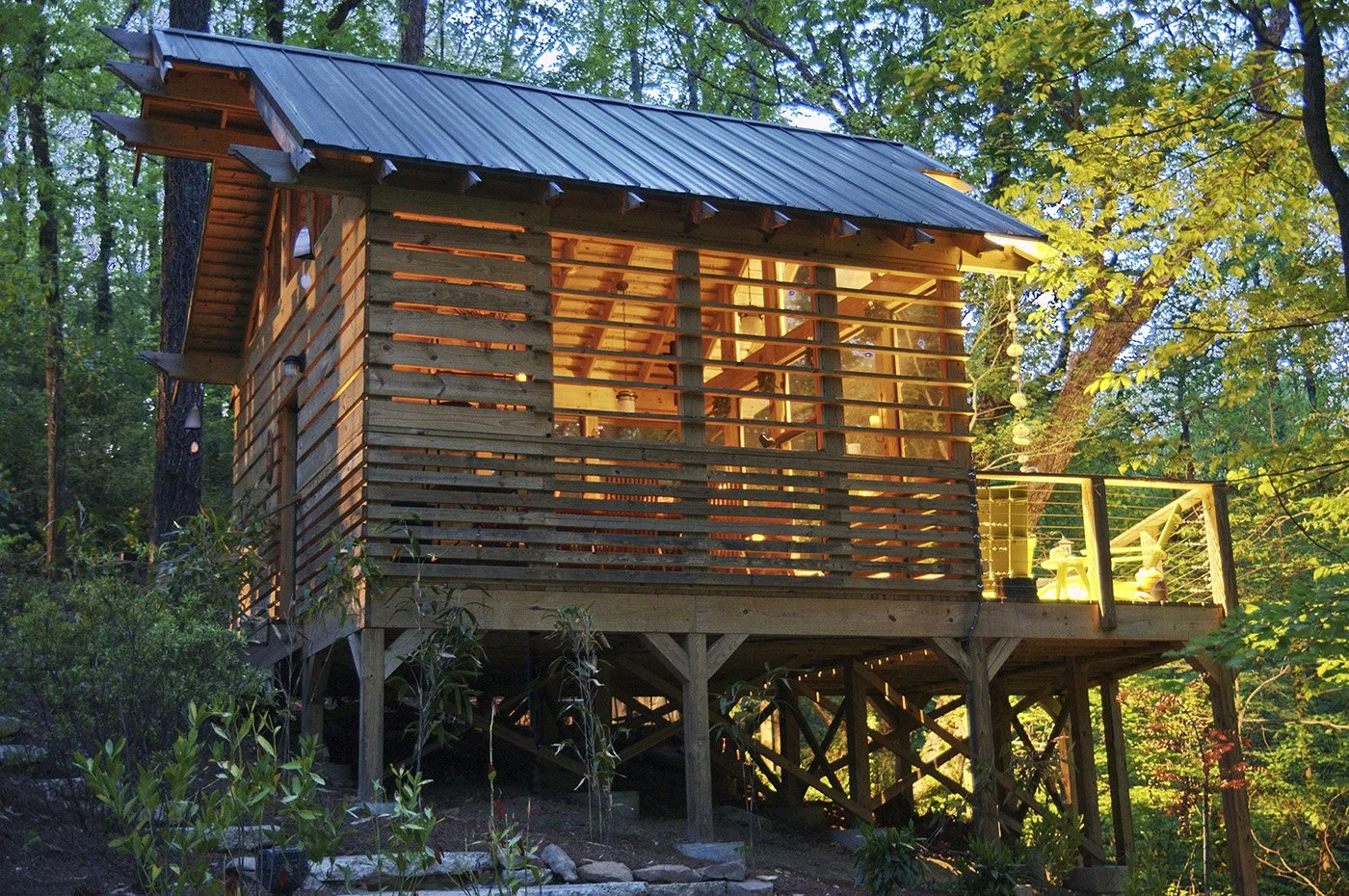 A wooden treehouse with a metal roof, built among trees with green foliage, illuminated from inside, featuring a balcony with string lights and outdoor furniture.