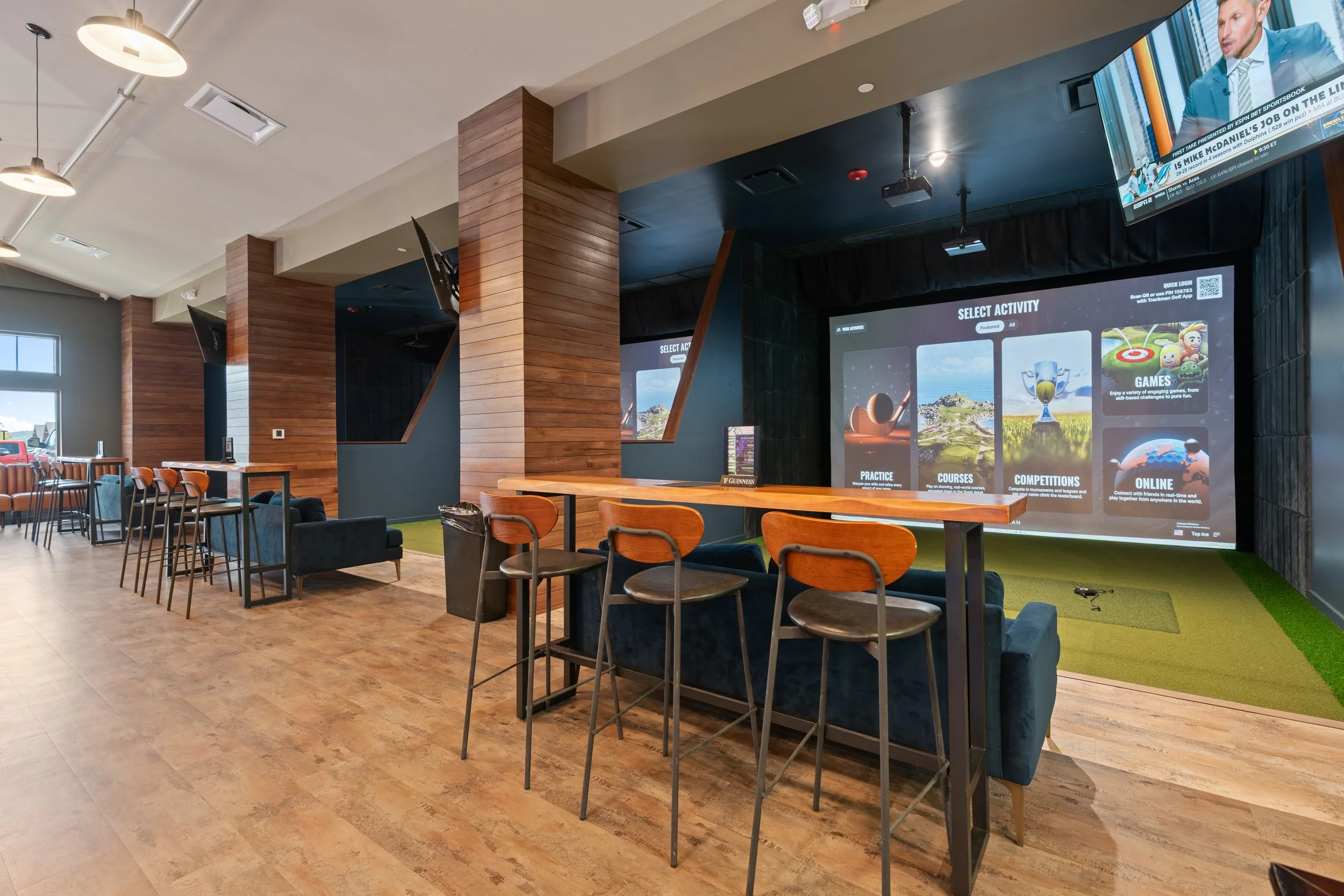 Interior of a modern entertainment venue with high tables and chairs, large video screens displaying gaming options, and a golf simulation area with artificial turf. project by B Group Architecture, Inc