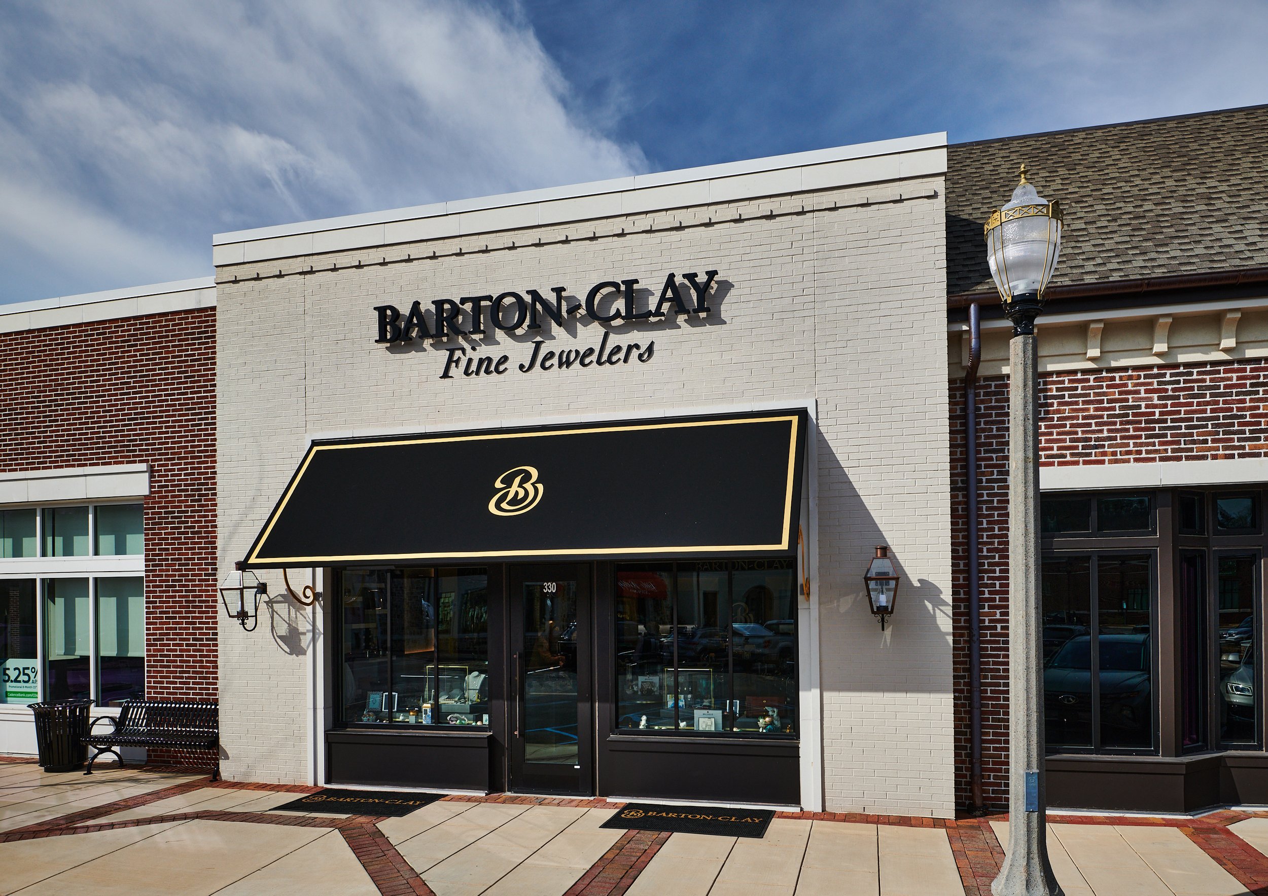 Front view of a jewelry store named Barton Clay Fine Jewelers, with a custom black and gold accented awning, glass display windows, and a painted cream brick exterior project by B Group Architecture, Inc