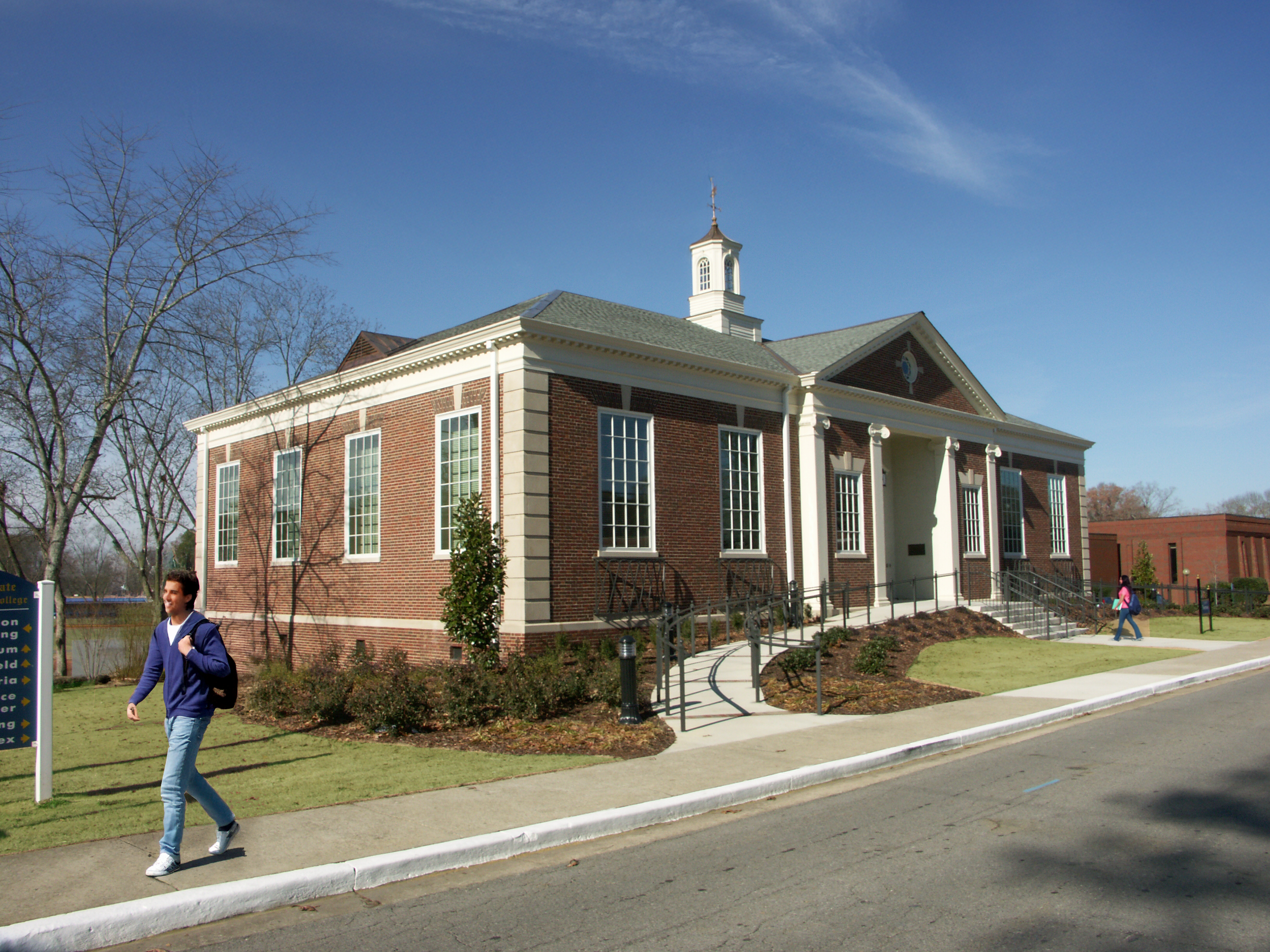 A brick school building with tall windows, white columns, and a small clock tower against a blue sky. Two students, one male and one female, walk on the sidewalk.