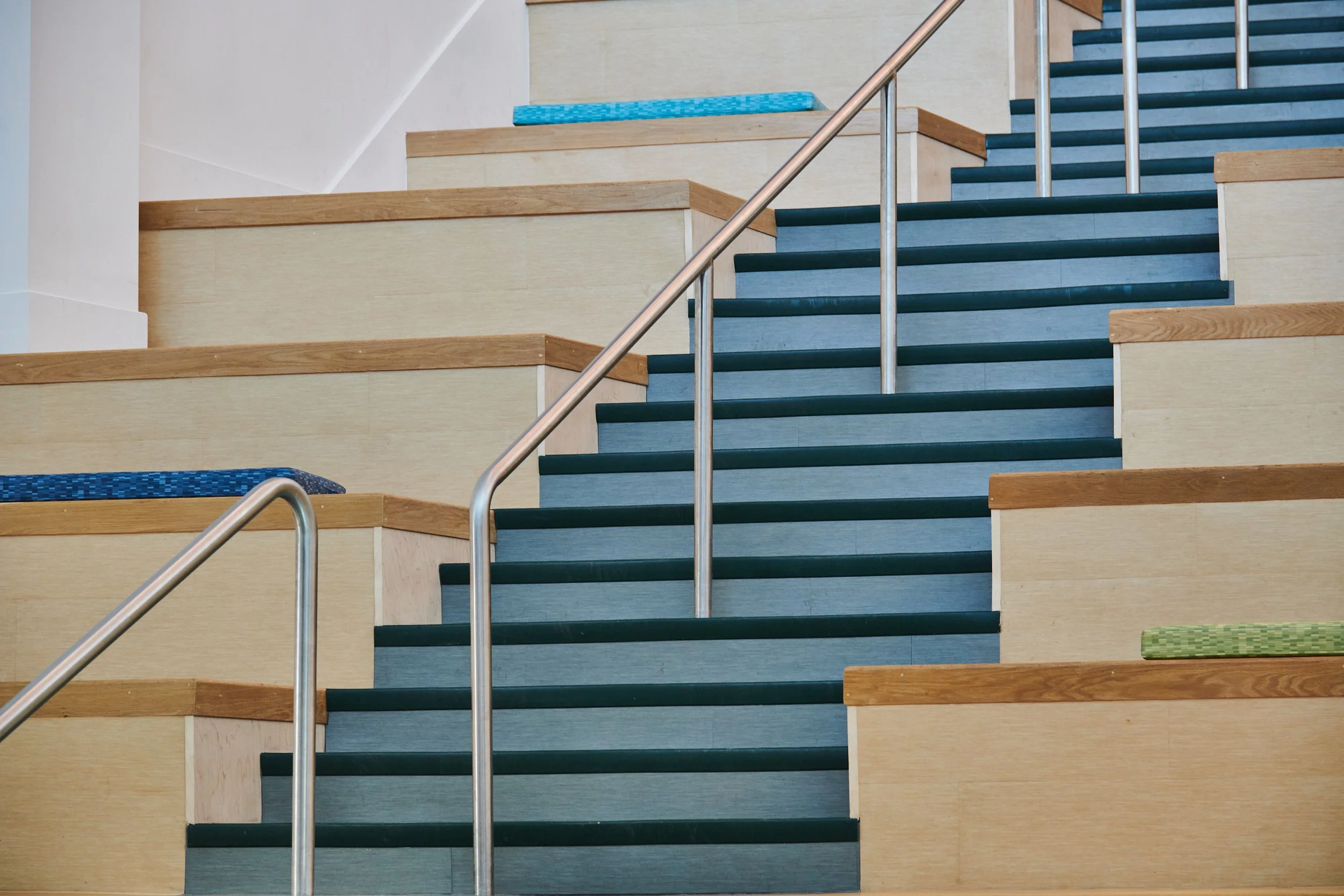 Indoor staircase with green steps, wooden edges, metal handrails, and colorful cushions on tiered high school commons seating- project by B Group Architecture, Inc.