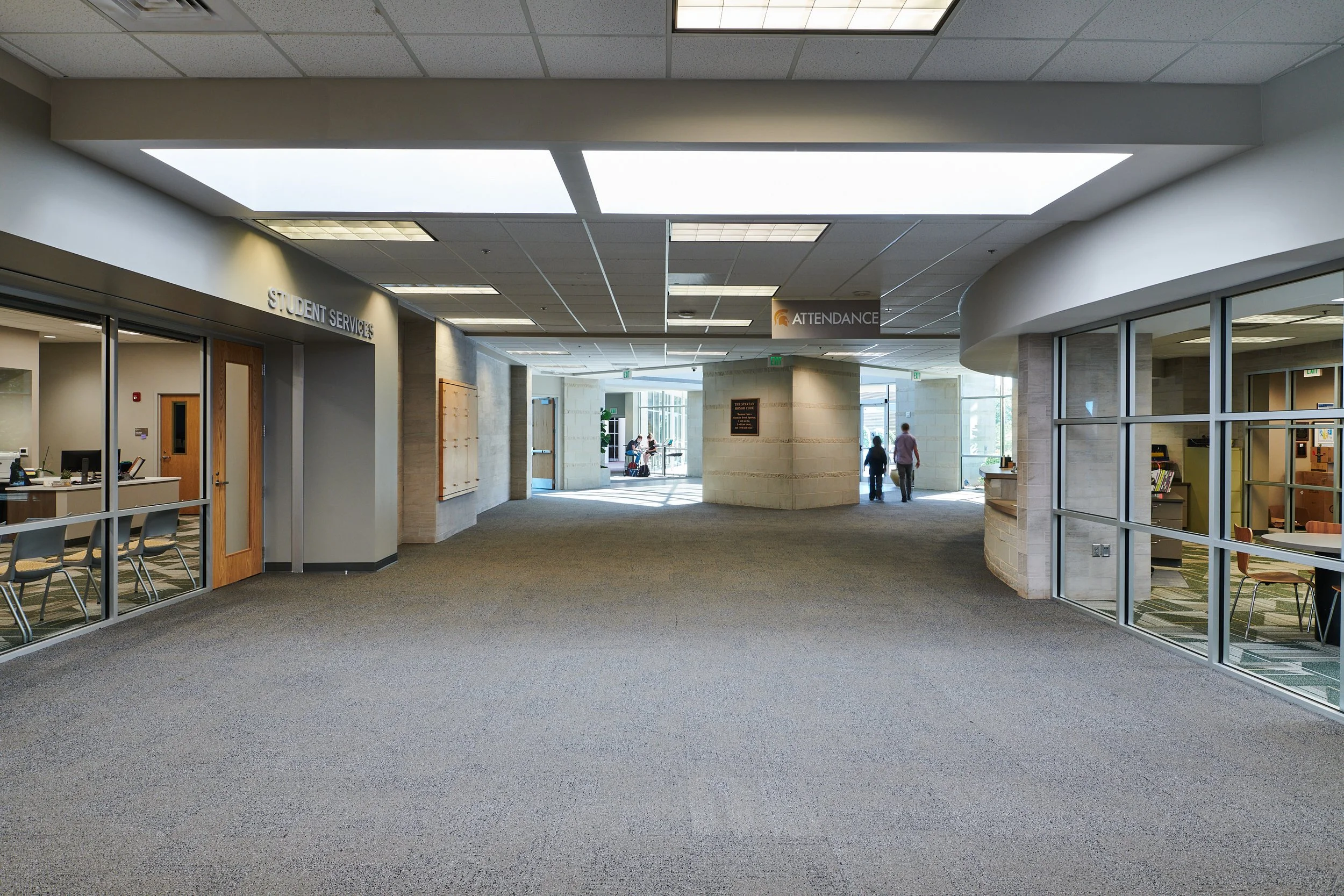 Interior of a modern lobby area, glass walls, and signs for Student Services and Attendance, with people walking and sitting in the background- project by B Group Architecture, Inc.